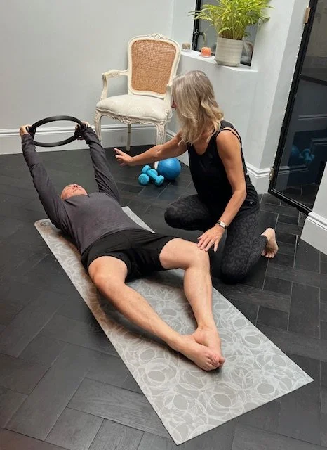 A woman in workout gear is leading an exercise session with two people. One person is lying on a yoga mat performing a leg raise, while another is lying on a Pilates reformer doing a similar exercise. The instructor is standing with arms outstretched, demonstrating the movement.