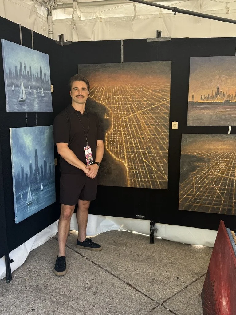 A man standing in an art display booth with paintings of cityscapes and aerial views of city streets at sunset or dusk.