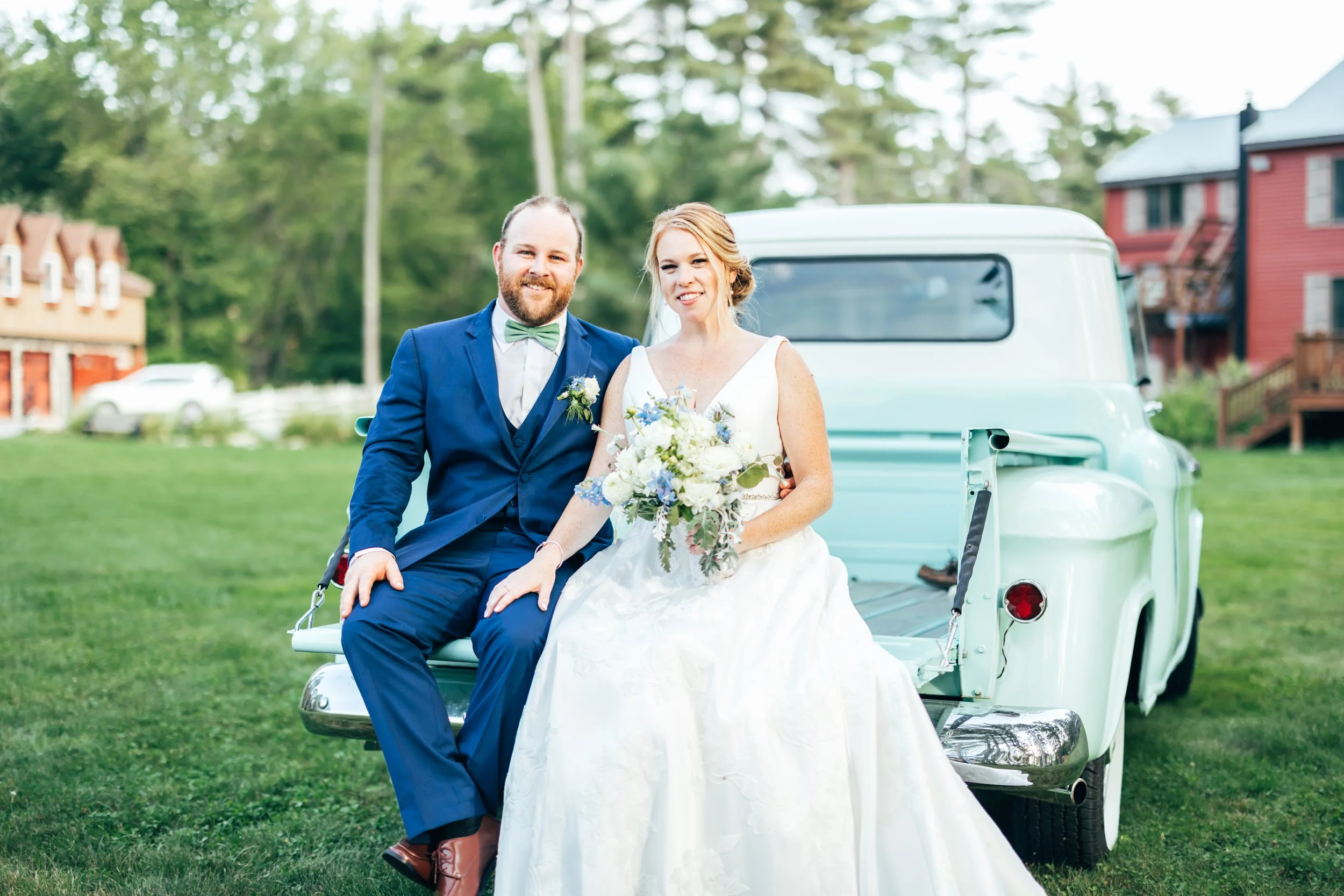 Wedding couple sitting on a vintage light blue truck outdoors, the bride in a white gown holding a bouquet, the groom in a blue suit with a green bow tie, in a grassy area with colorful houses and trees in the background.