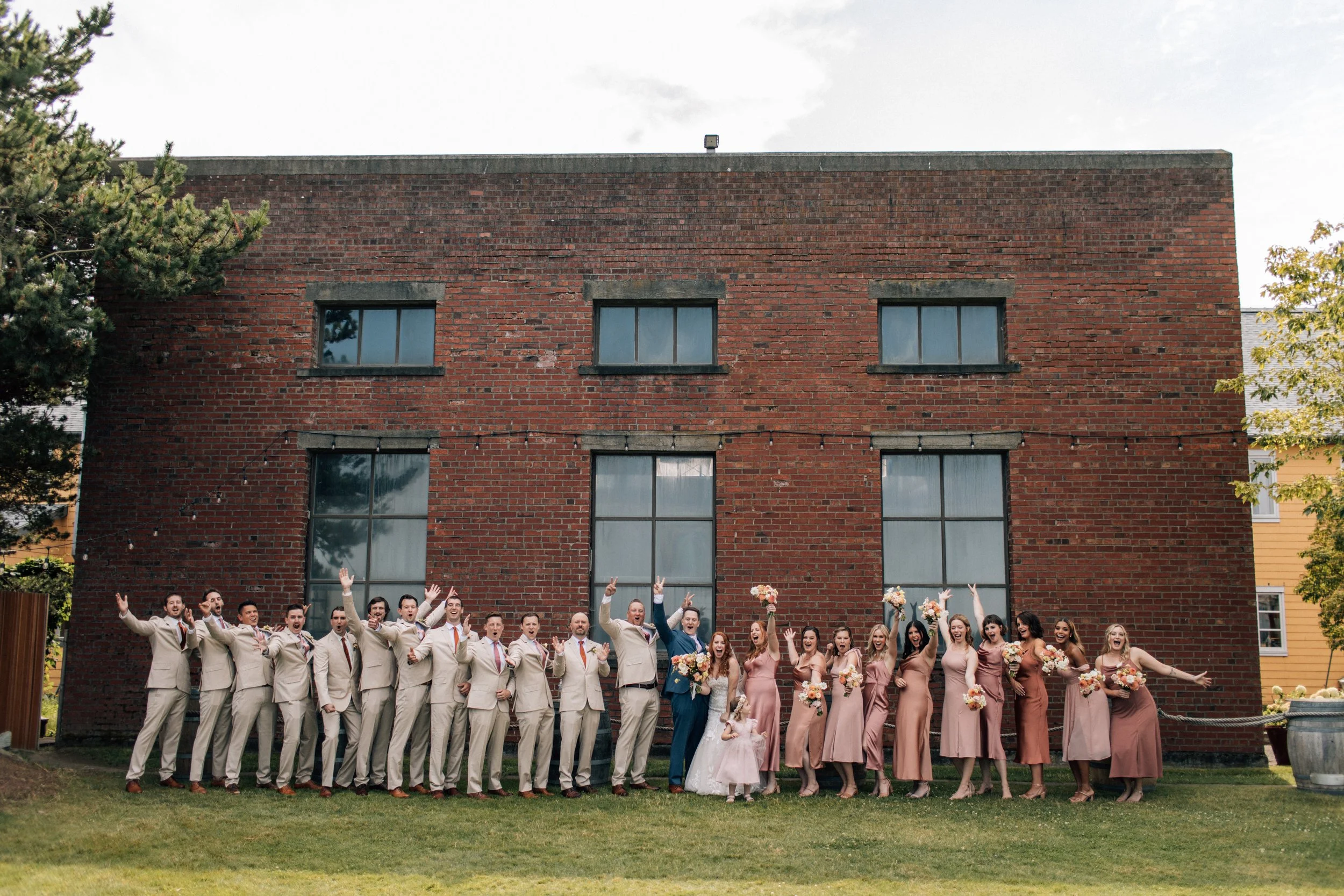 Wedding party standing in front of a brick building, group photo with bridesmaids and groomsmen, bride and groom in center, holding bouquets, celebratory poses