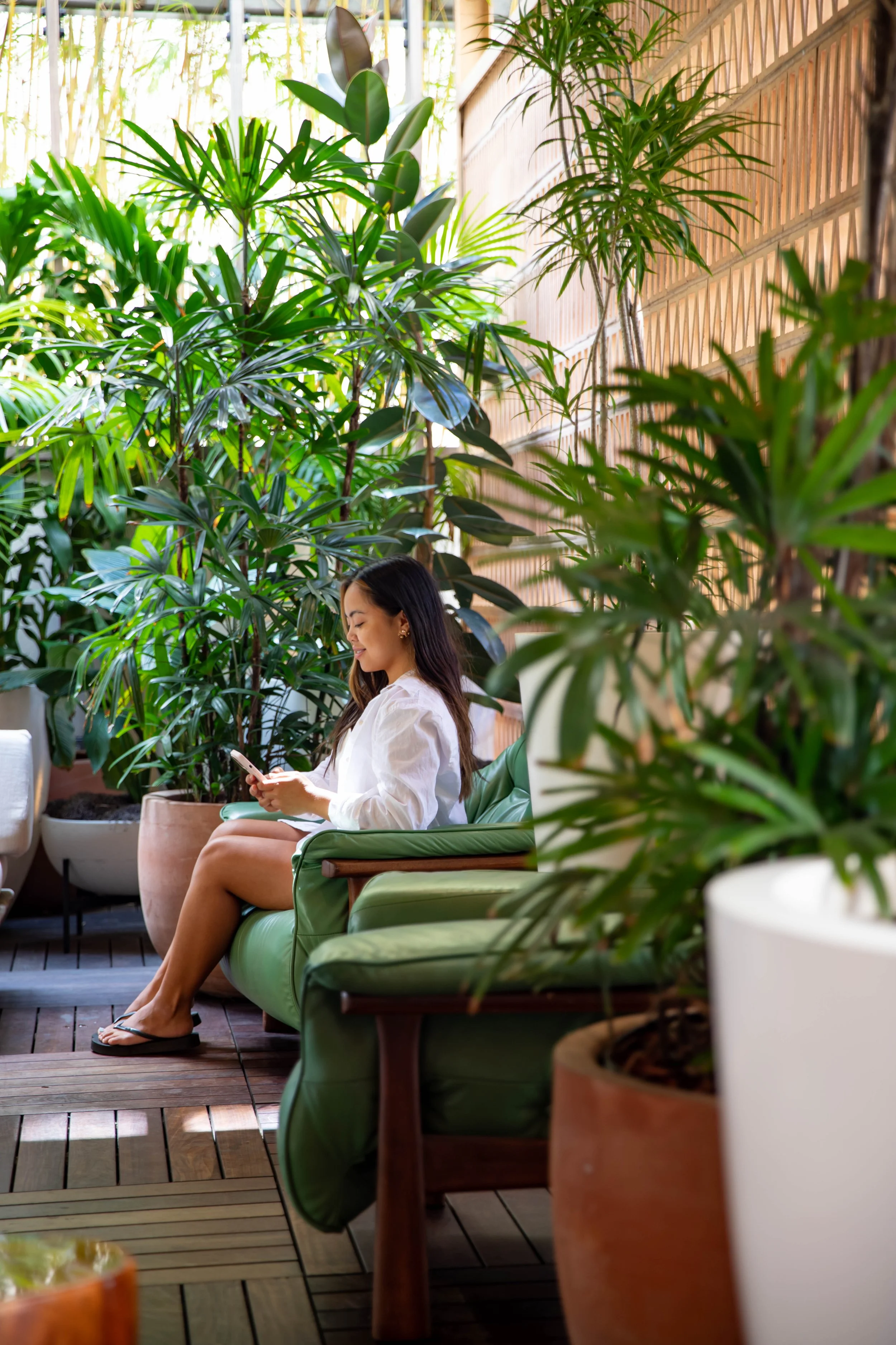 A woman sitting on a green lounge chair in a lush indoor garden, looking at her phone with potted plants surrounding her and sunlight filtering through windows.