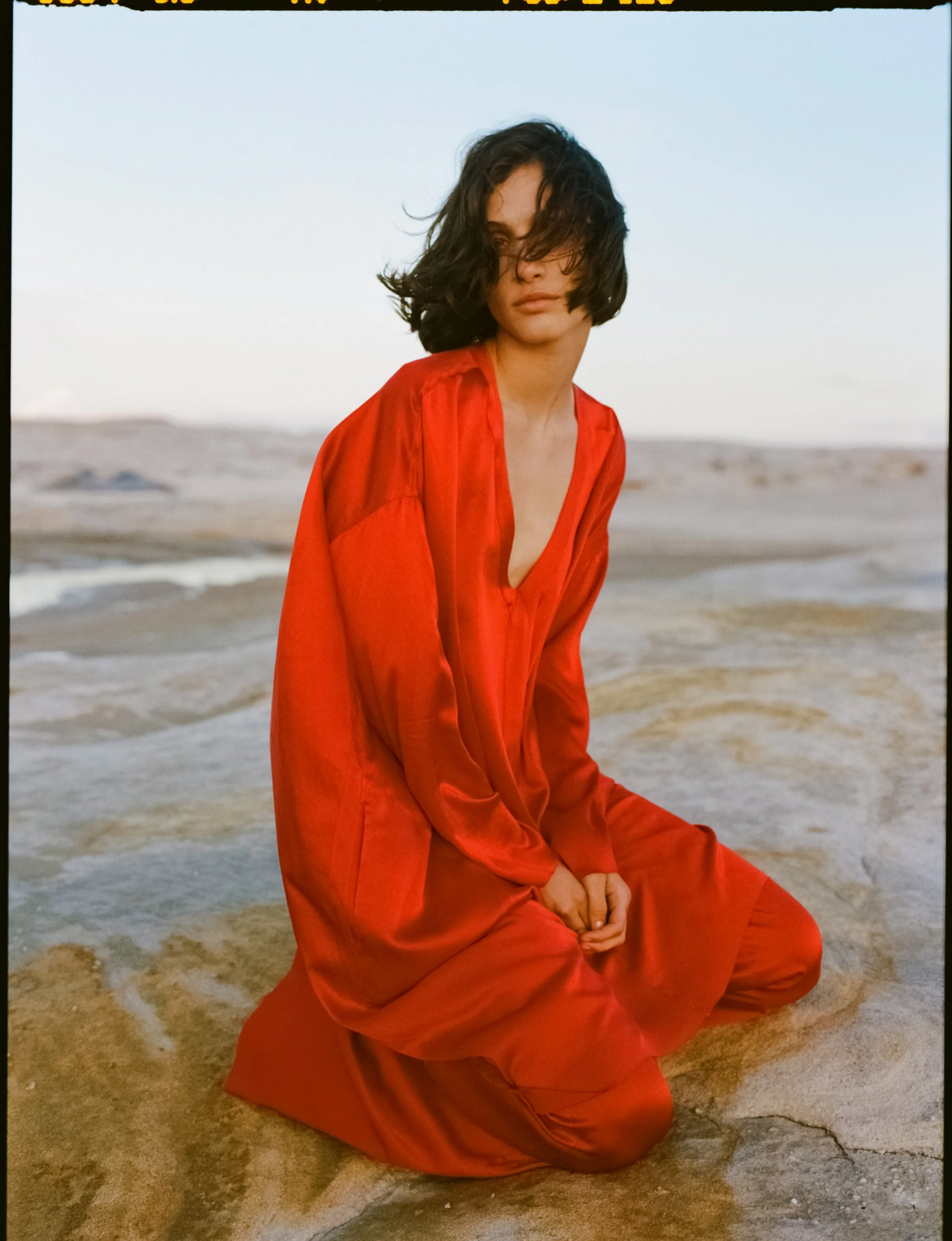 A woman in a loose red satin dress kneels on a sandy beach with ocean waves in the background, her hair partially covering her face.