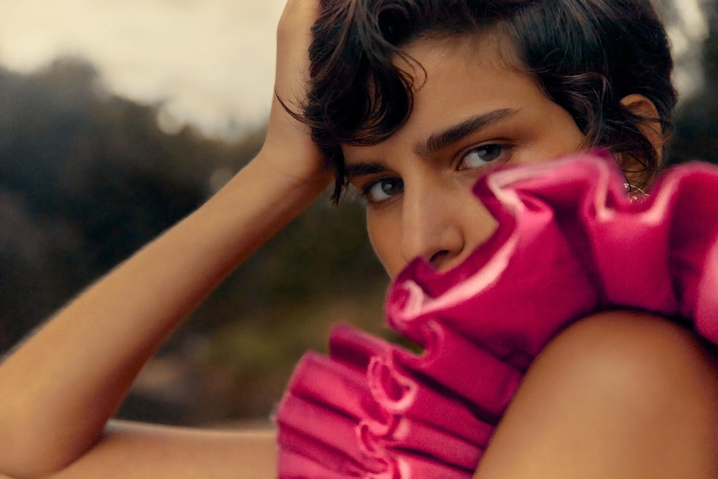 A young woman with short curly brown hair, posing outdoors with her hand resting on her head, wearing a bright pink, ruched top, and looking into the camera with a contemplative expression.