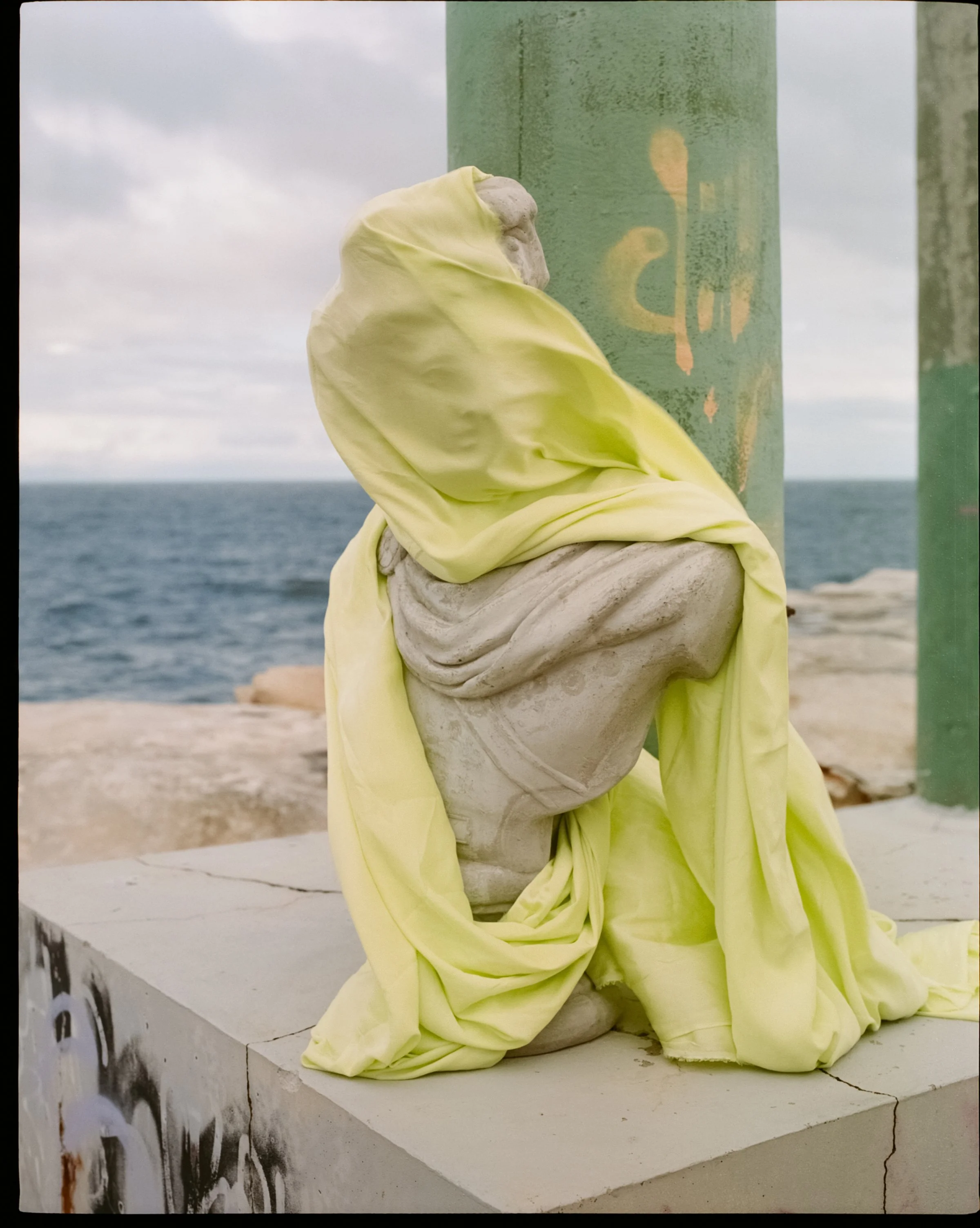 A weathered sculpture of a person wrapped in a yellow cloth, seated near the ocean on a concrete surface with a green pole beside it, under an overcast sky.