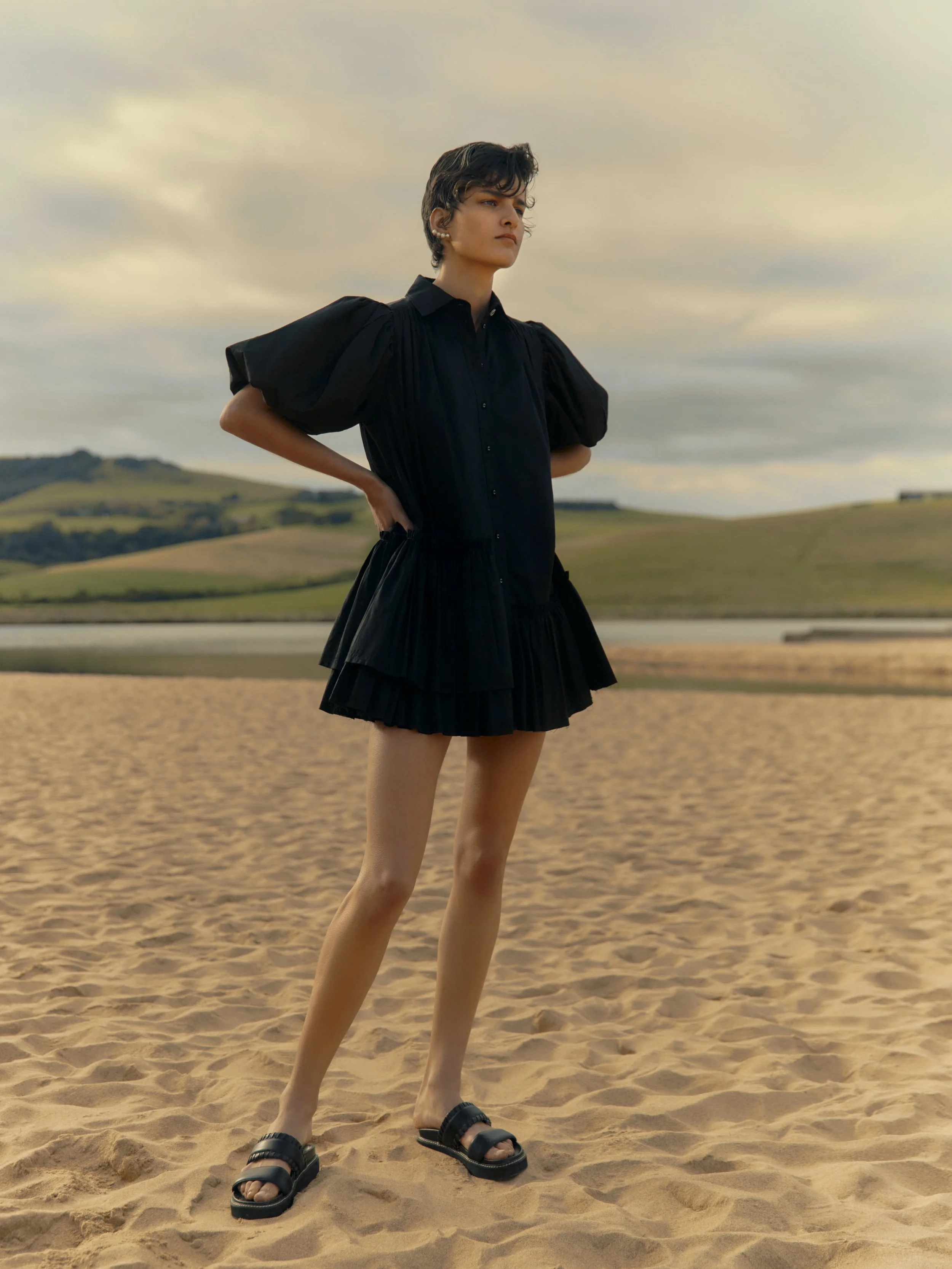 A young woman standing on a sandy beach with hills and a cloudy sky in the background, wearing a black dress with puffed sleeves and black slide sandals.