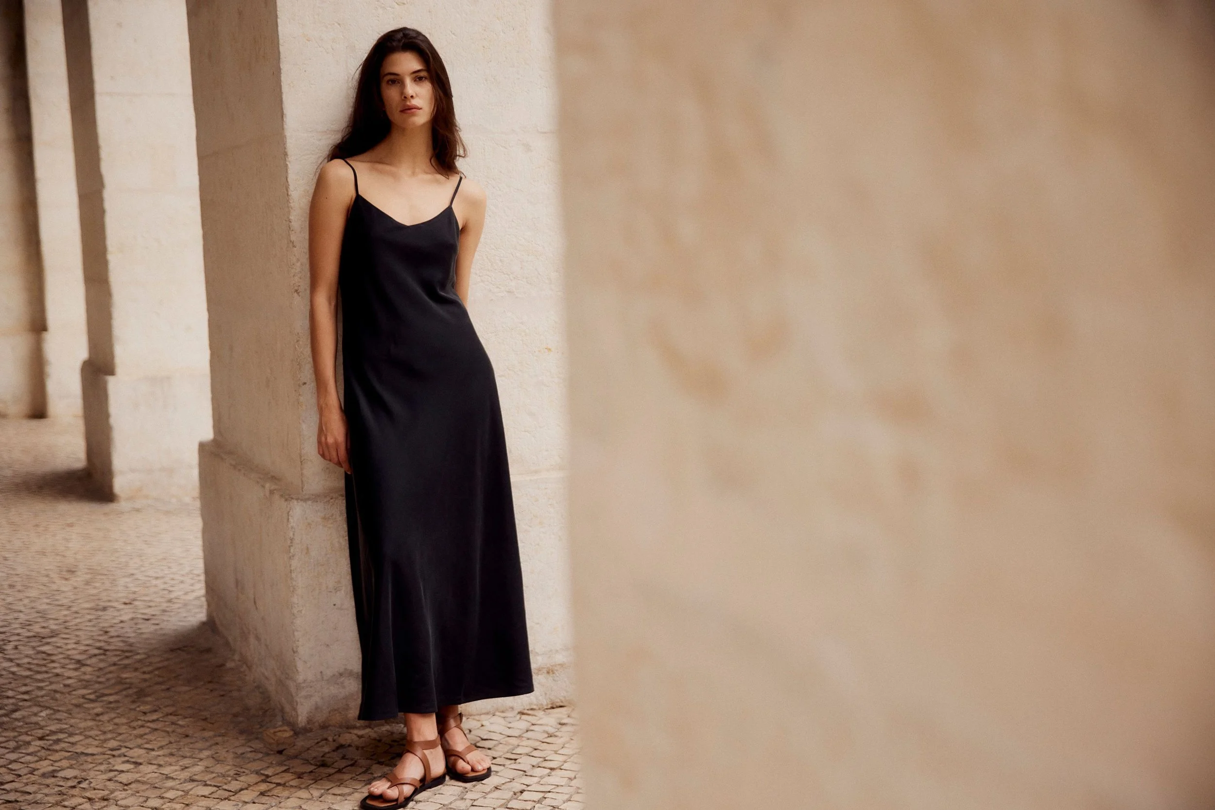 A young woman with long dark hair leaning against a beige stone wall, wearing a black slip dress and sandals, in an outdoor setting.