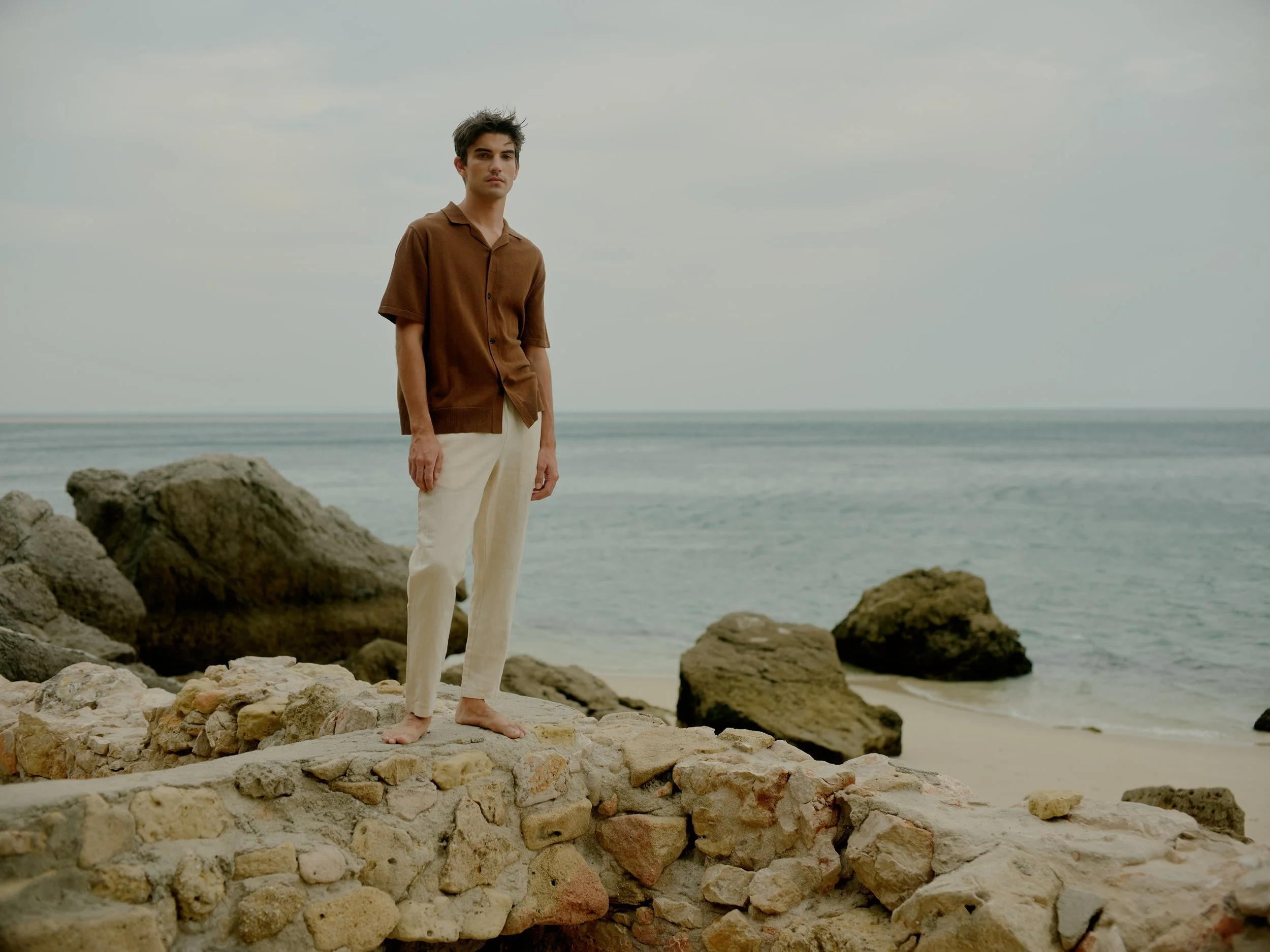 A man standing barefoot on a rocky beach, wearing a brown short-sleeved shirt and cream-colored pants, with the ocean and cloudy sky in the background.