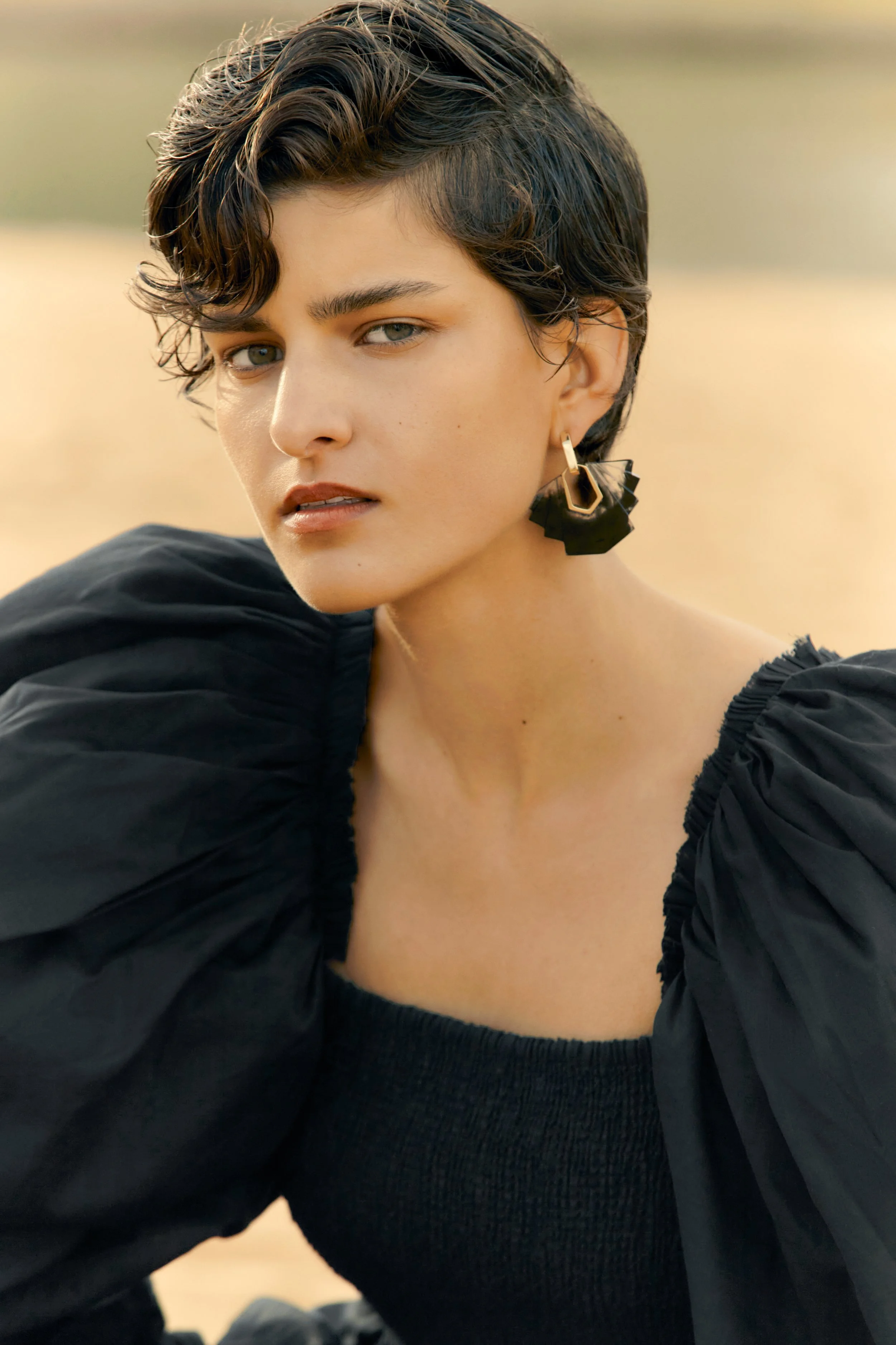 A woman with short dark curly hair, wearing a black puffed sleeve top and large geometric black and gold earrings, poses outdoors with a blurred natural background.