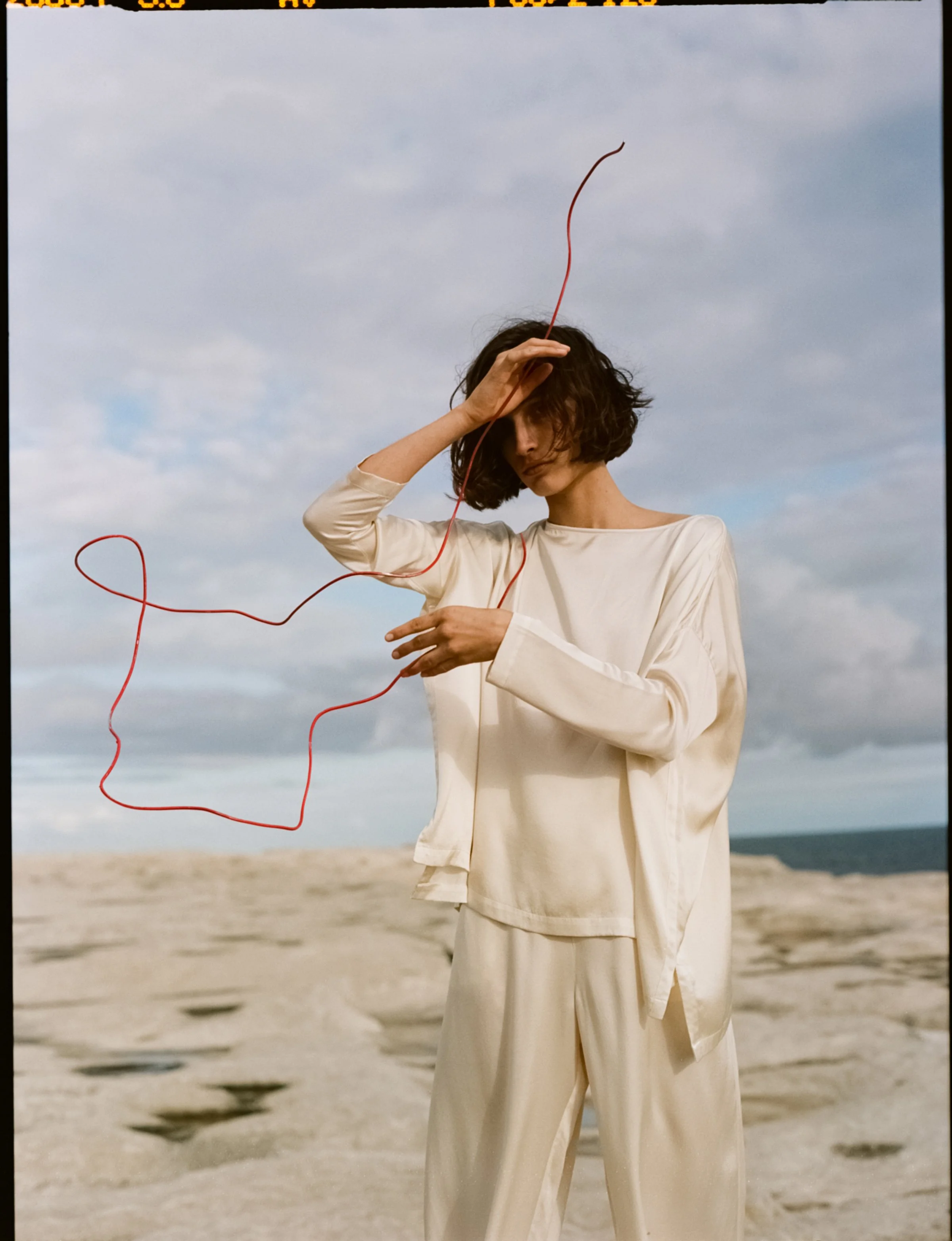 A woman dressed in cream-colored clothing standing on a sandy beach, holding a red wire shaped like a face, with a cloudy sky in the background.