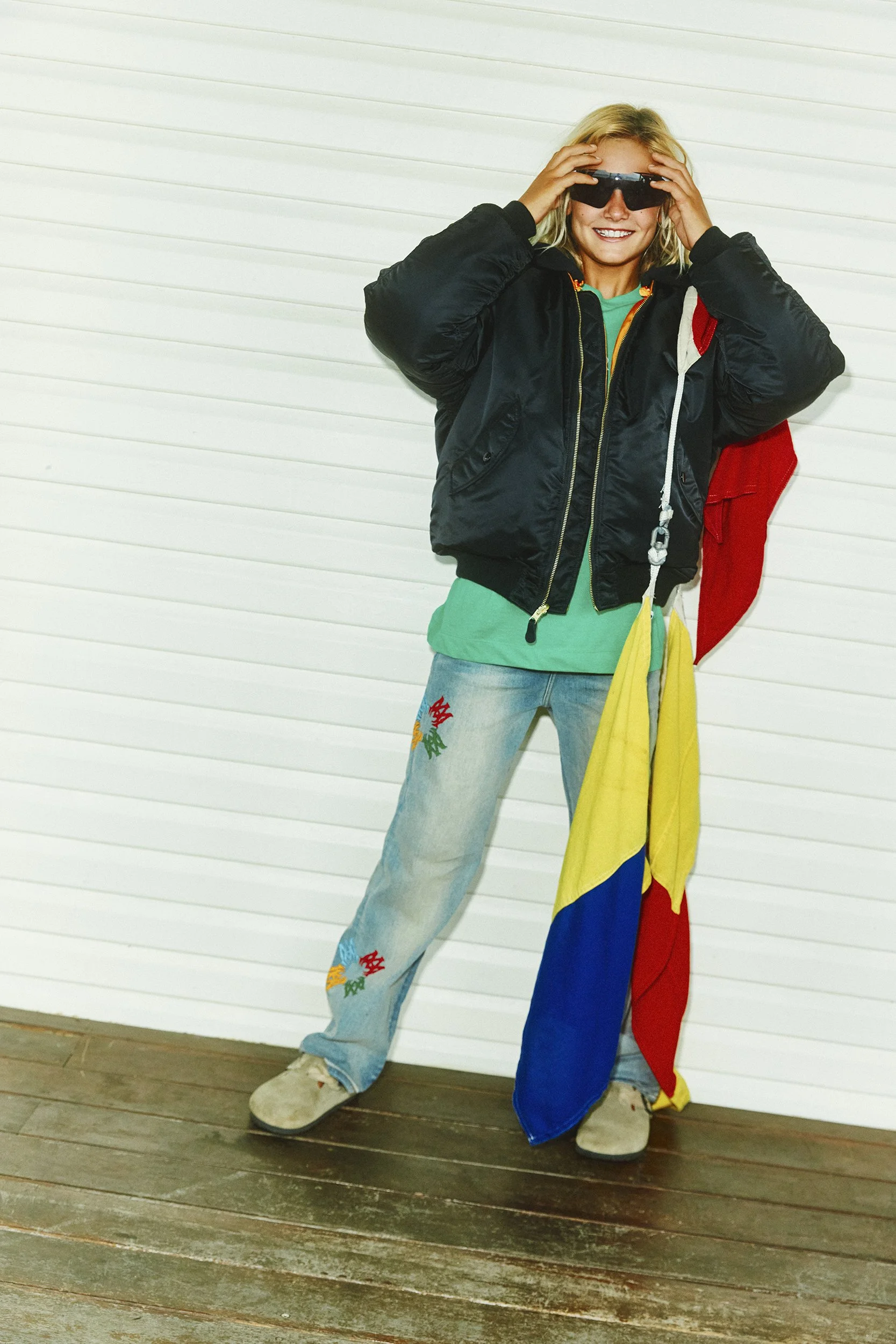 A woman standing in front of a white garage door, wearing sunglasses, a black jacket, blue jeans with colorful embroidery, and beige shoes. She is holding her head with both hands and smiling.