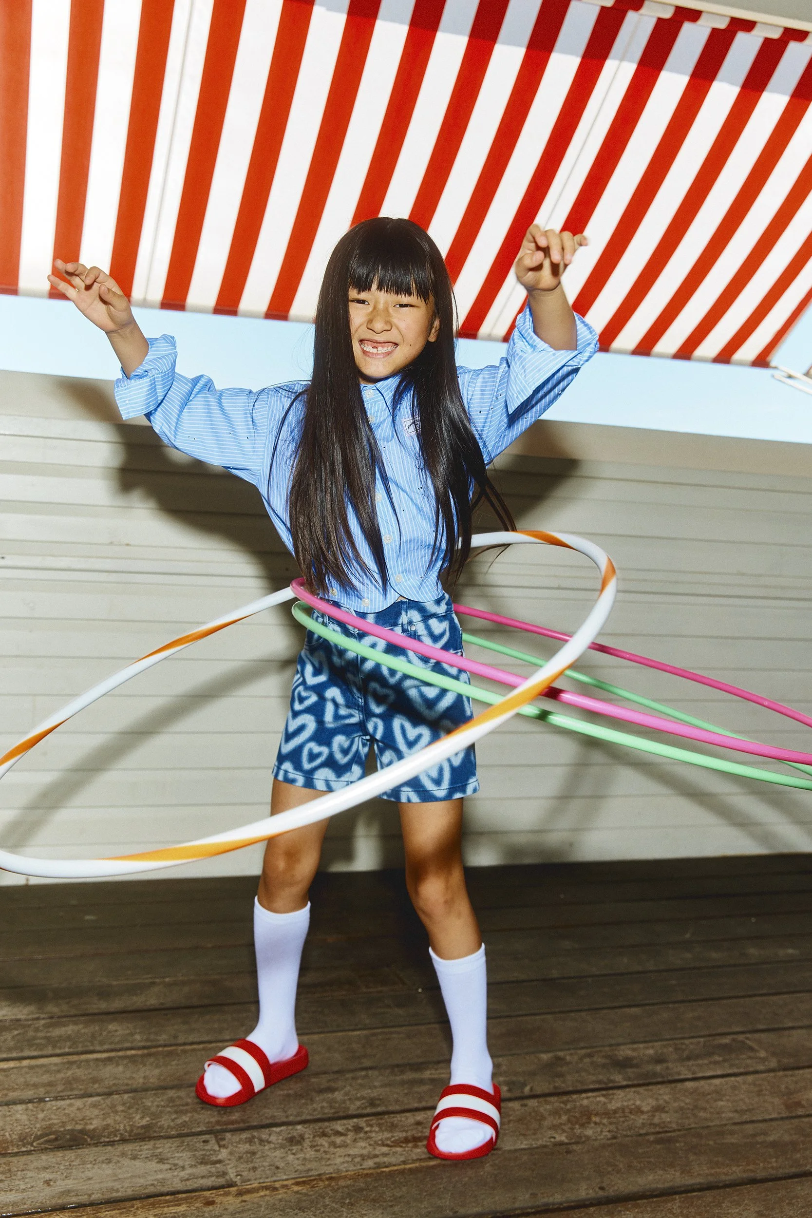 A young girl with long black hair smiling and posing with multiple hula hoops around her waist and arms, wearing a blue patterned shorts, a blue shirt, white knee-high socks, and red and white slides, standing on a wooden deck with a red and white st
