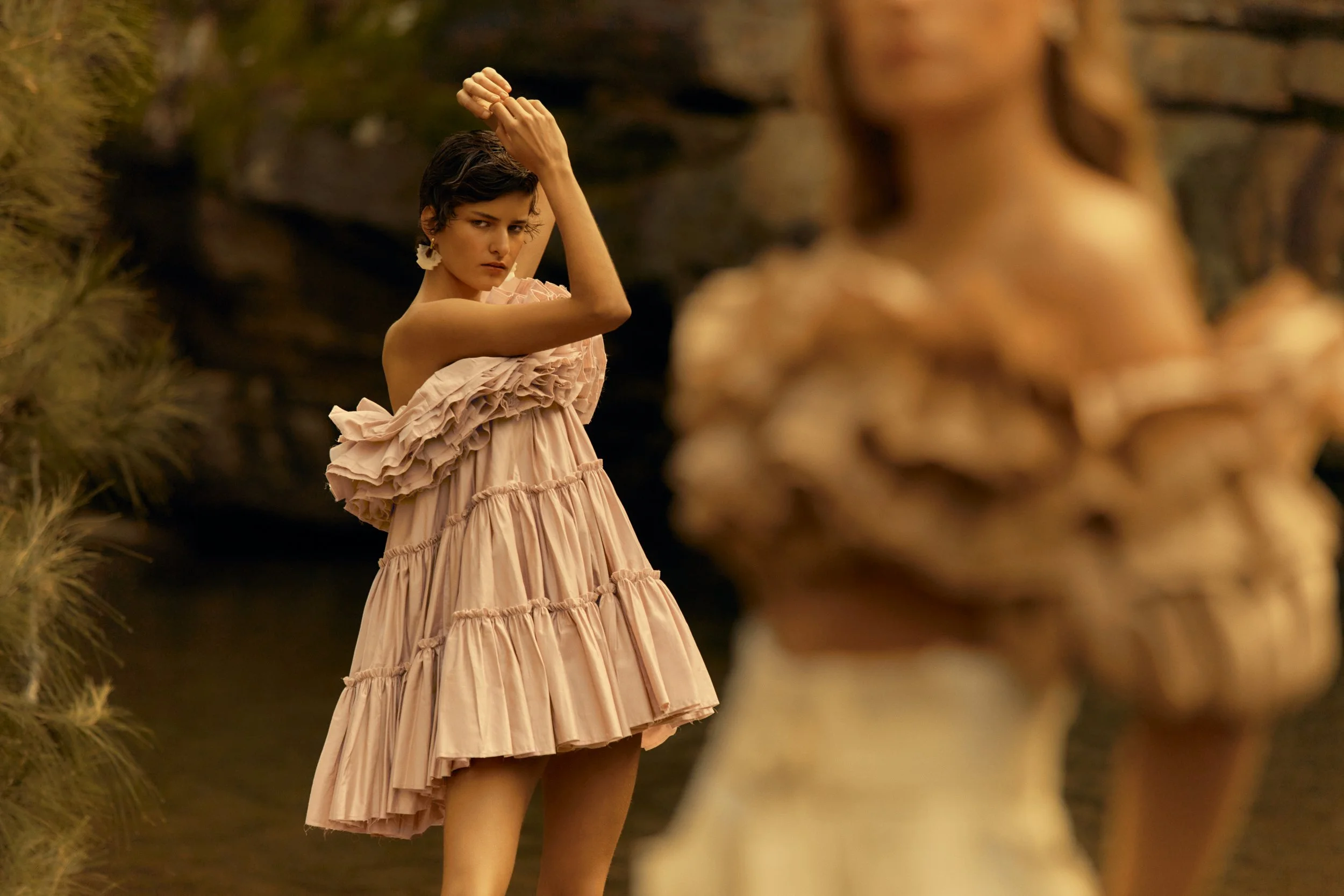 A woman in a pink ruffled dress standing outdoors near a rocky formation, with her arm raised above her head. Another woman in the foreground is blurred.