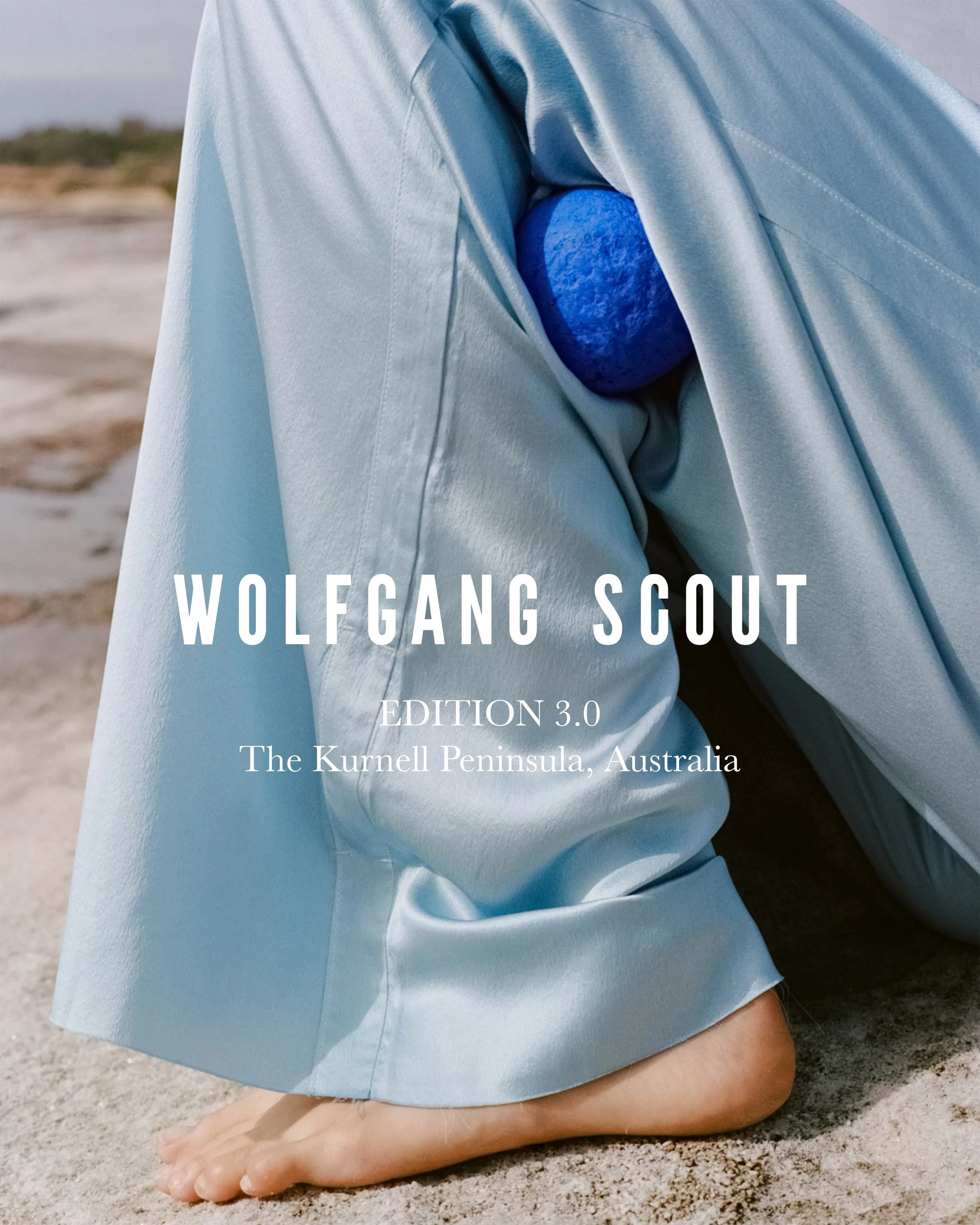 Close-up of a person in a light blue robe kneeling on sandy ground, with a blue stone in the robe's pocket. The background is a beach scene. Text overlay reads 'Wolfgang Scout Edition 3.0 The Kurnell Peninsula, Australia.'