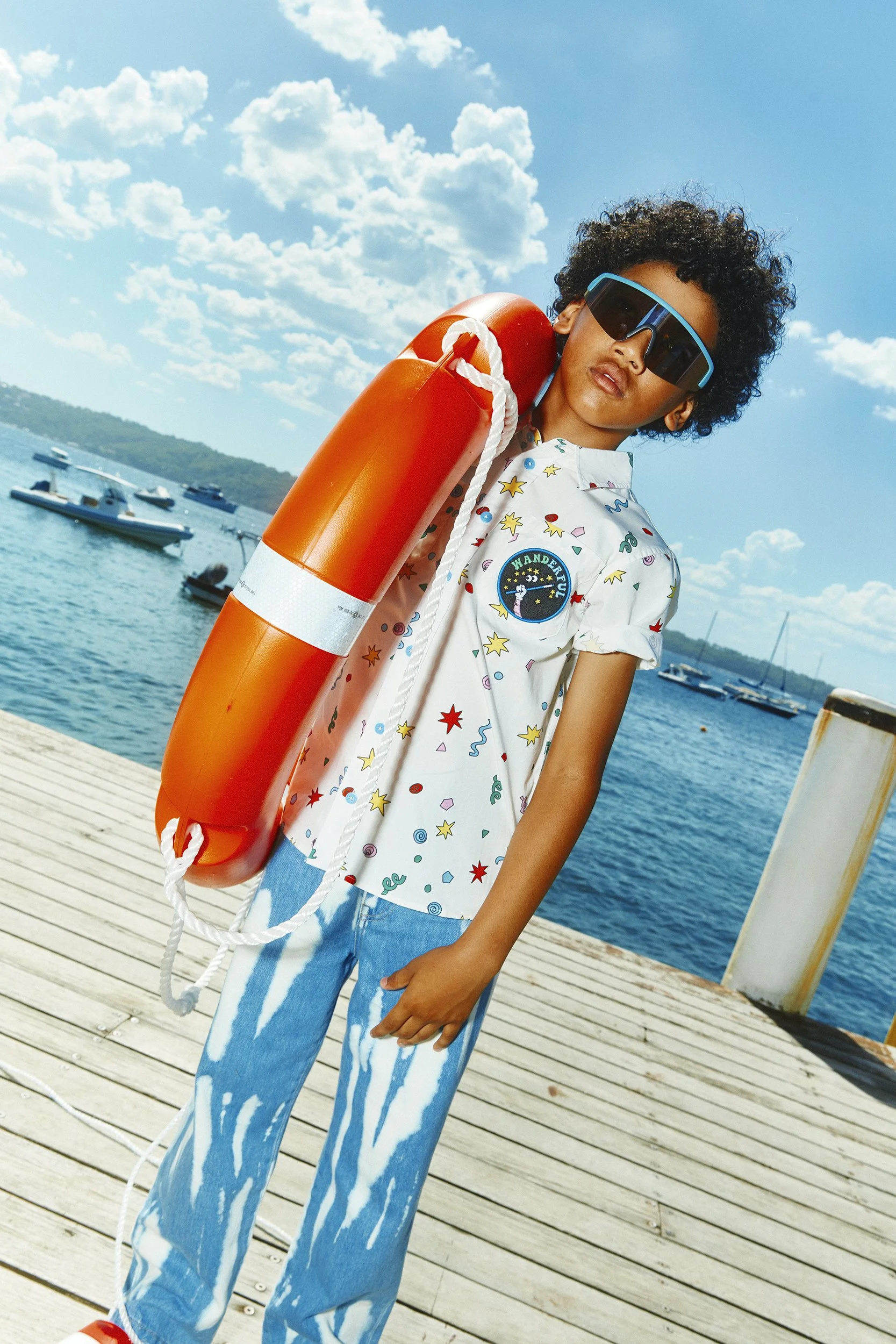 A person standing on a wooden dock by the water, holding an orange life-saving ring. They are wearing large sunglasses, a white short-sleeved shirt with colorful patterns, and blue tie-dye pants. The background shows boats on the water under a partly