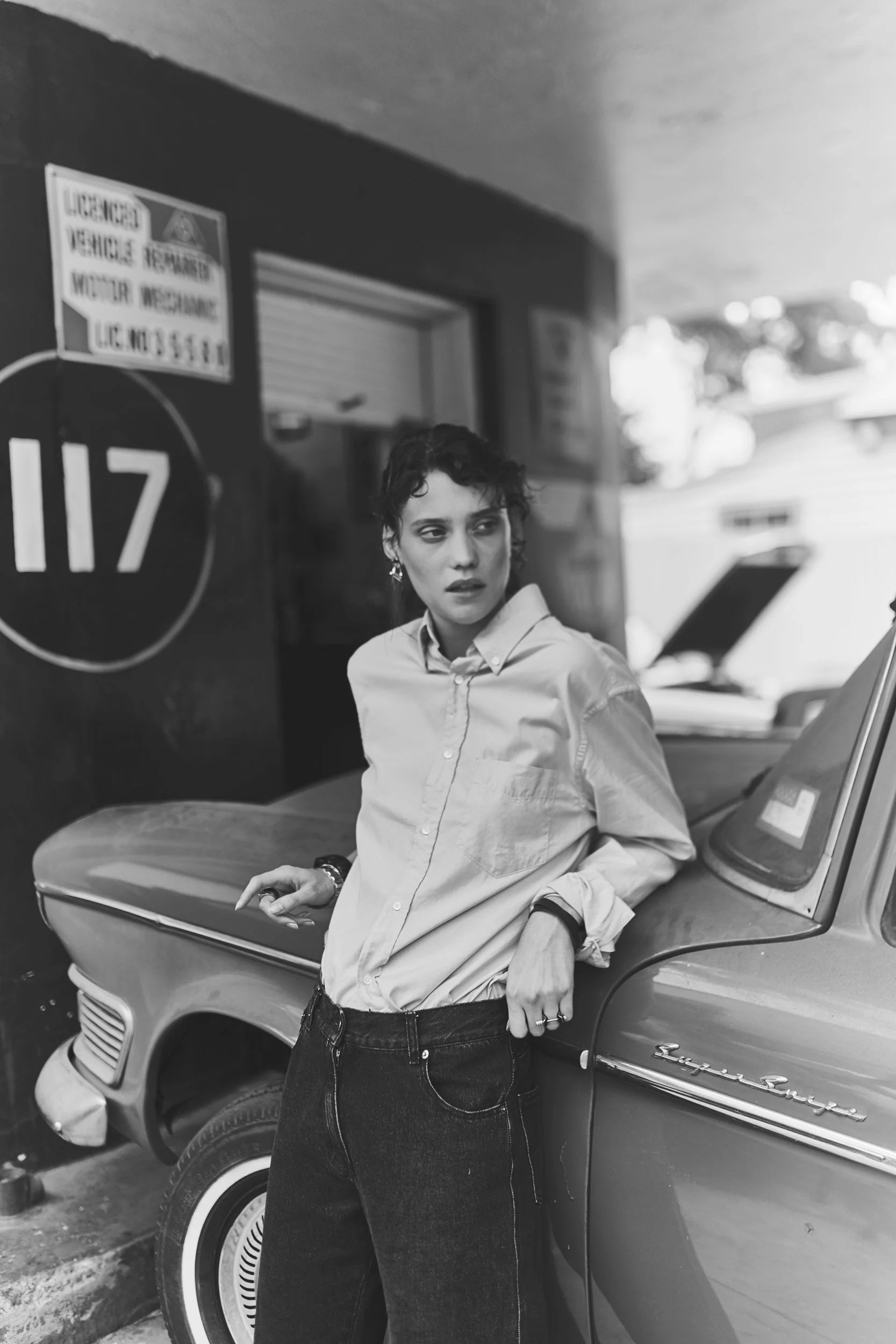 A woman with short curly hair, wearing a light-colored button-up shirt and dark jeans, leans against a vintage car with a sign and house numbers in the background.