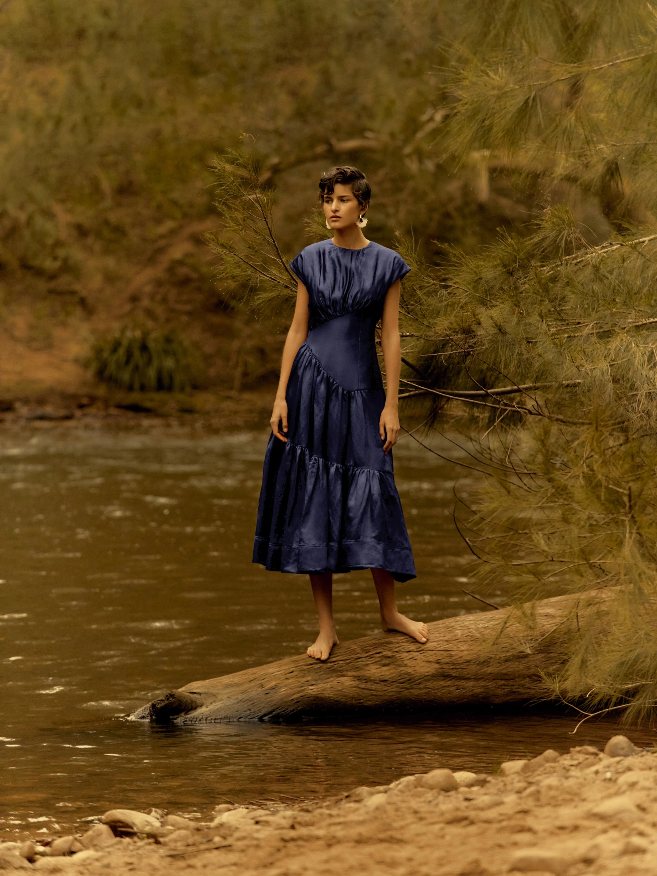 A woman in a dark blue dress standing barefoot on a log in a river surrounded by trees.