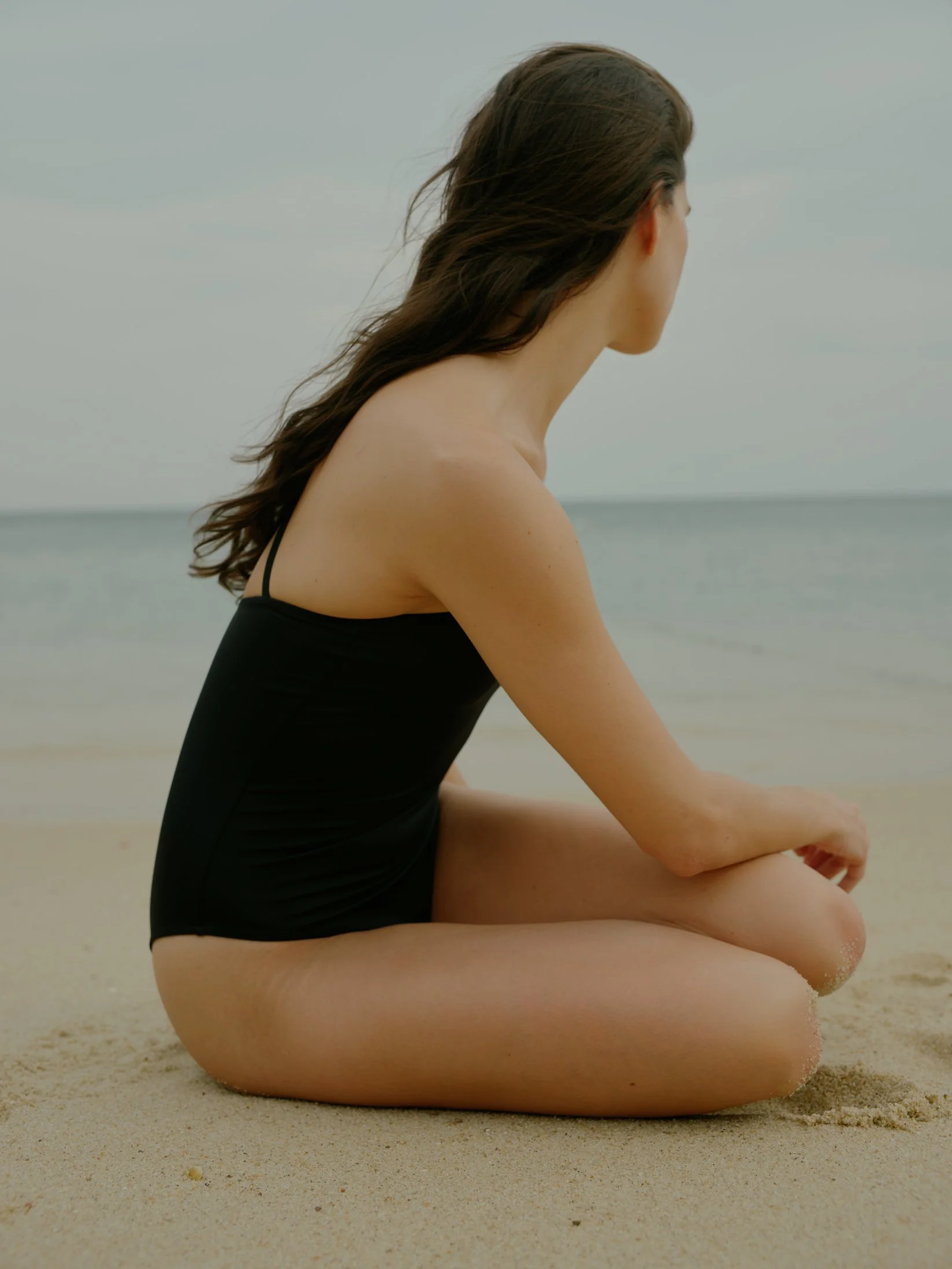 A woman with long brown hair sitting on the sandy beach in a black swimsuit, facing away from the camera, near the water.