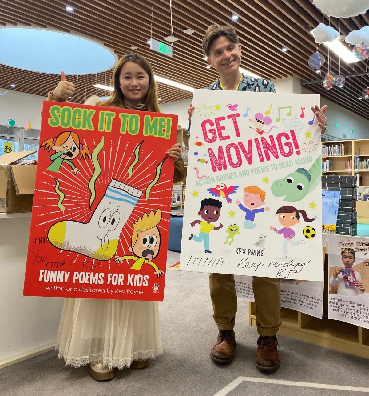 Kev Payne and a female Chinese teacher hold up printed out book covers that read: sock it to me, funny poems for kids and 'get moving, action rhymes and poems to read aloud' both written by Kev Payne