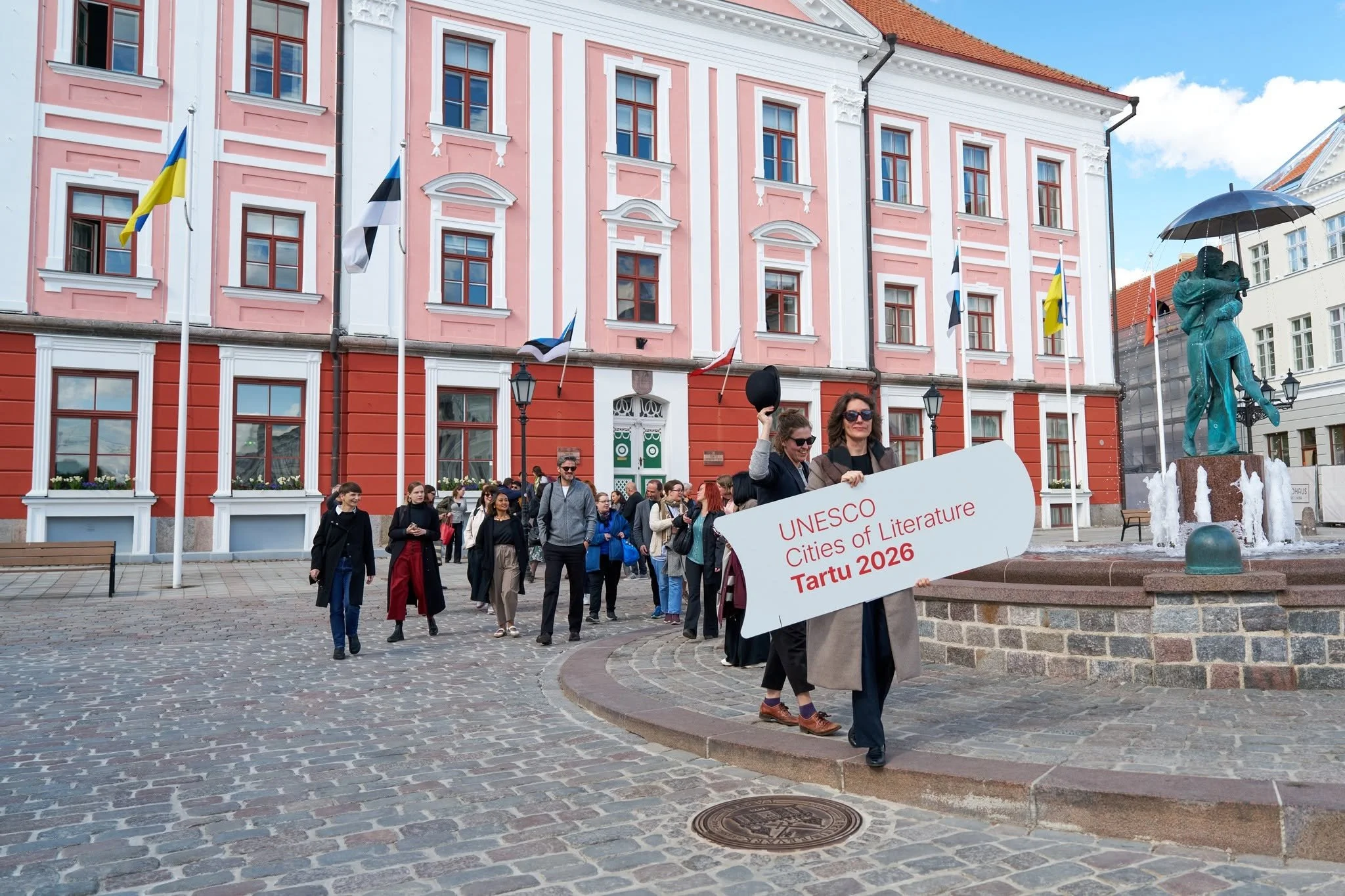 A woman holds a 'UNESCO Cities of Literature Tartu 2026' sign and directs a group of visitors across an old courtyard in Tartu, Estonia.