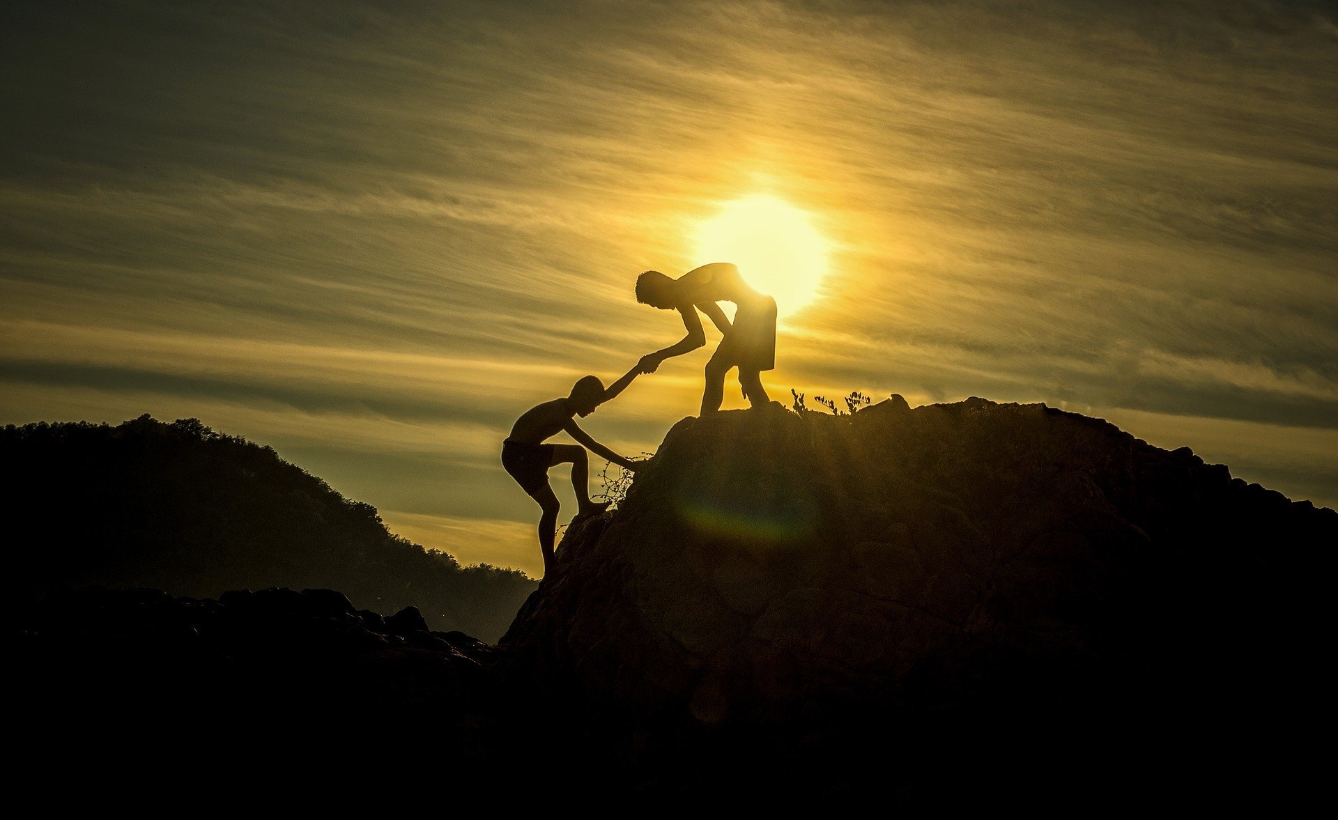 A person helps another person climb up a hill, silhouetted against the setting sun