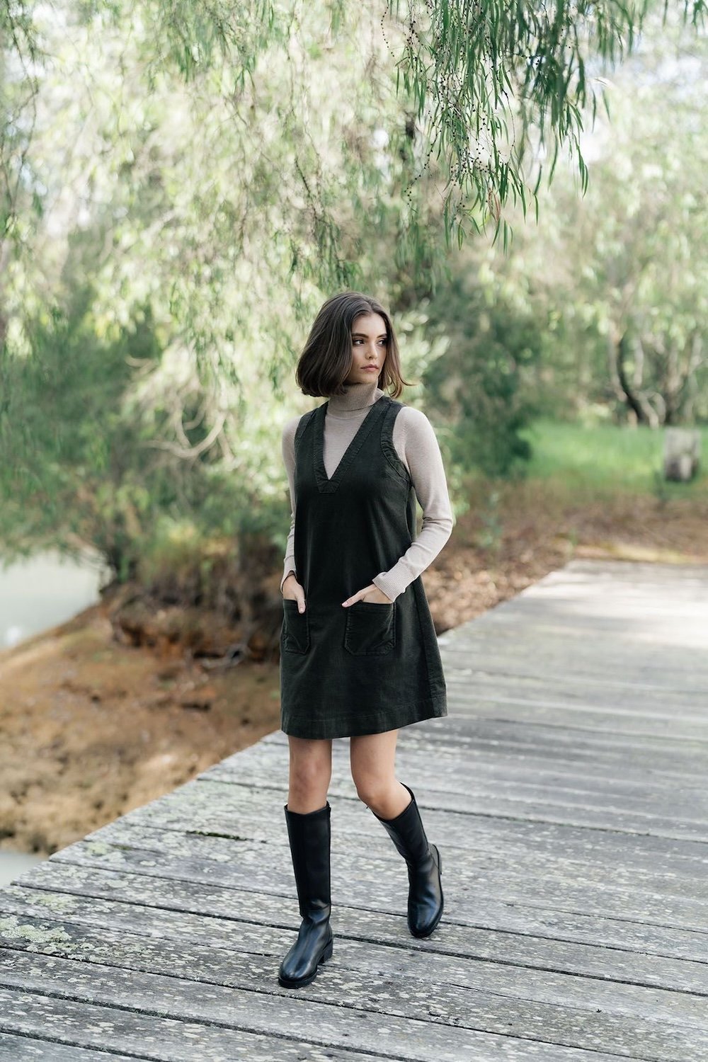 Girl wearing moss green cord a line dress with deep vee neck and finishing mid thigh.