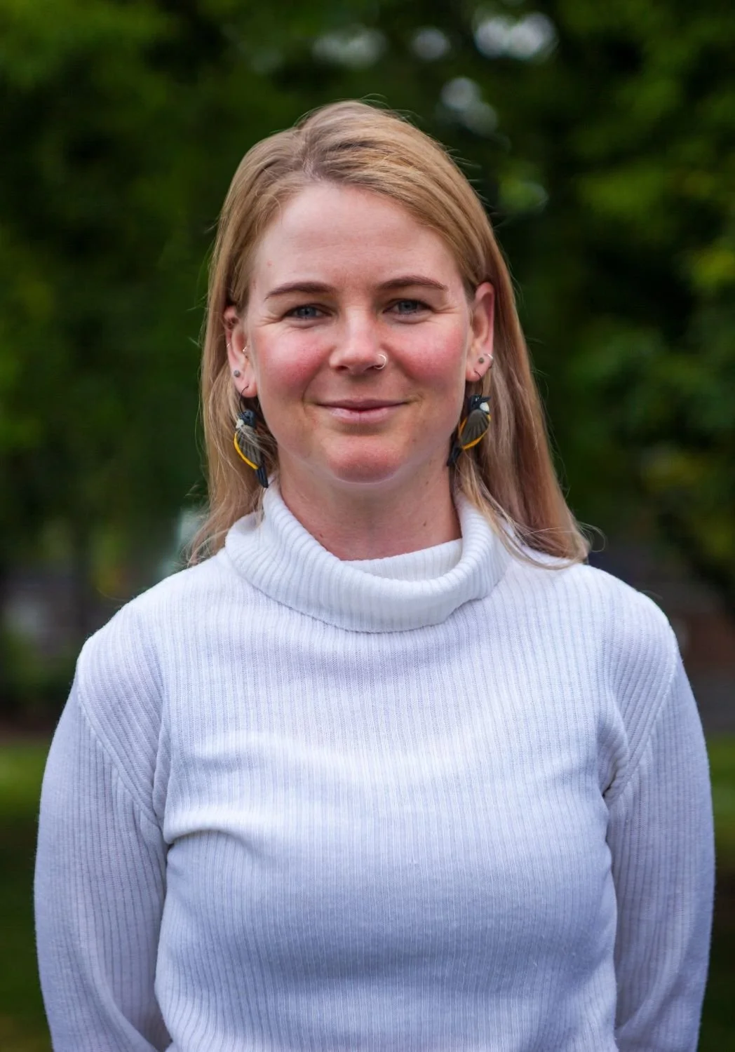 Close-up of a woman with reddish-blonde hair, wearing a white turtleneck sweater, smiling outdoors with a green, blurry background.