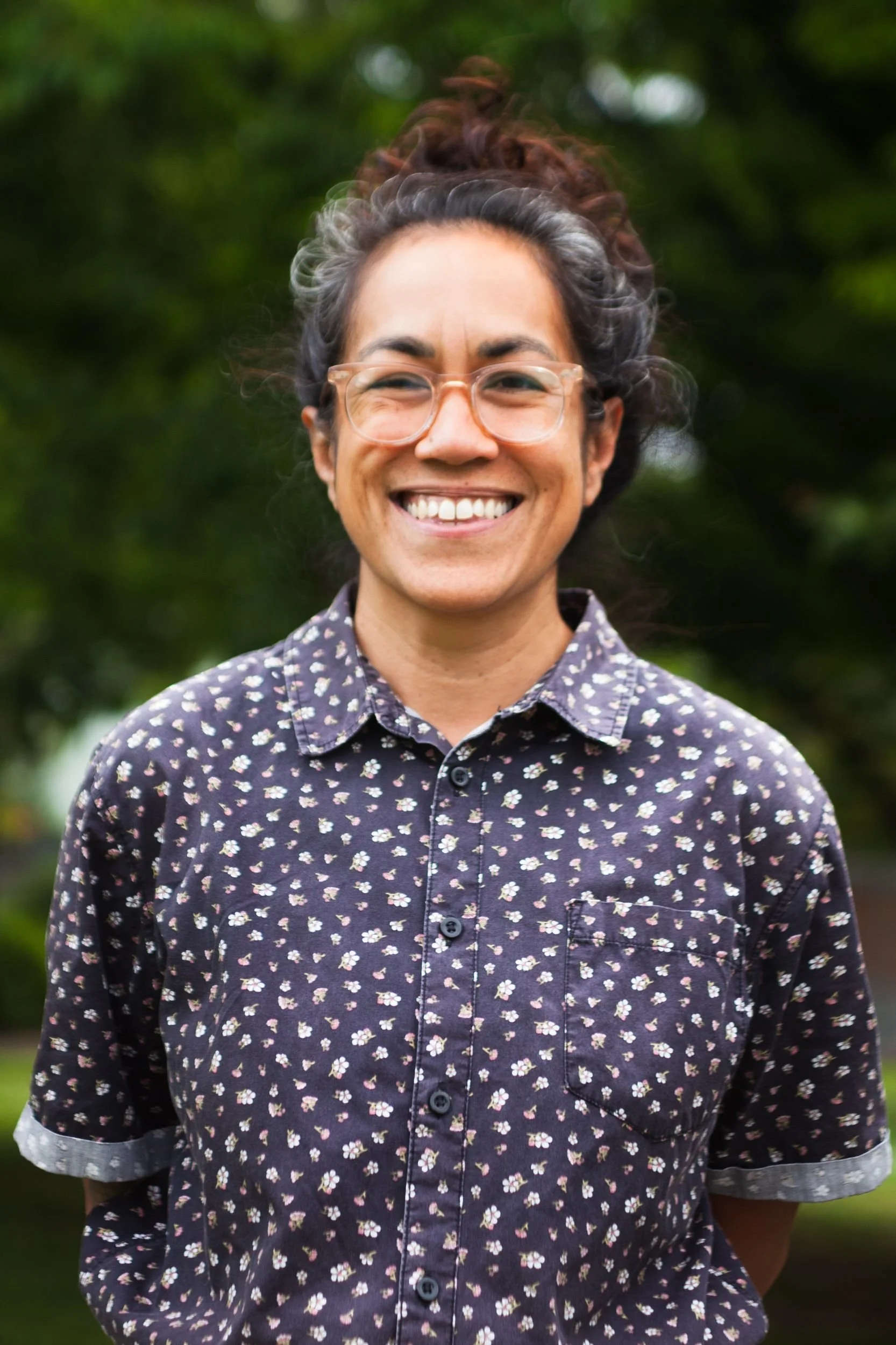 A woman with glasses and curly hair tied up, smiling outdoors in front of green trees, wearing a dark button-up shirt with small floral patterns.