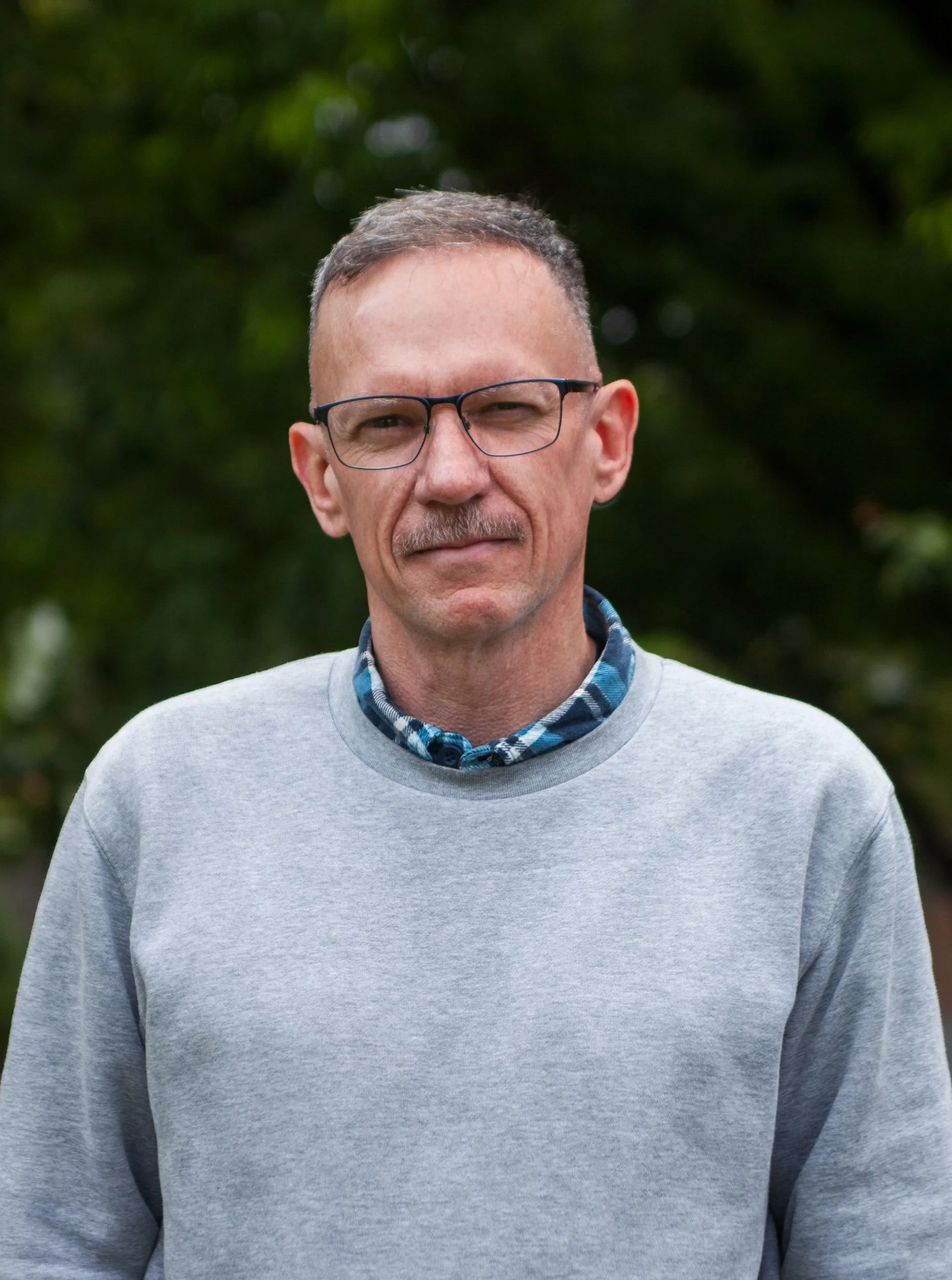 A smiling man with short gray hair, glasses, and a light mustache, wearing a gray sweatshirt over a blue and black plaid shirt, standing outdoors in front of green trees.