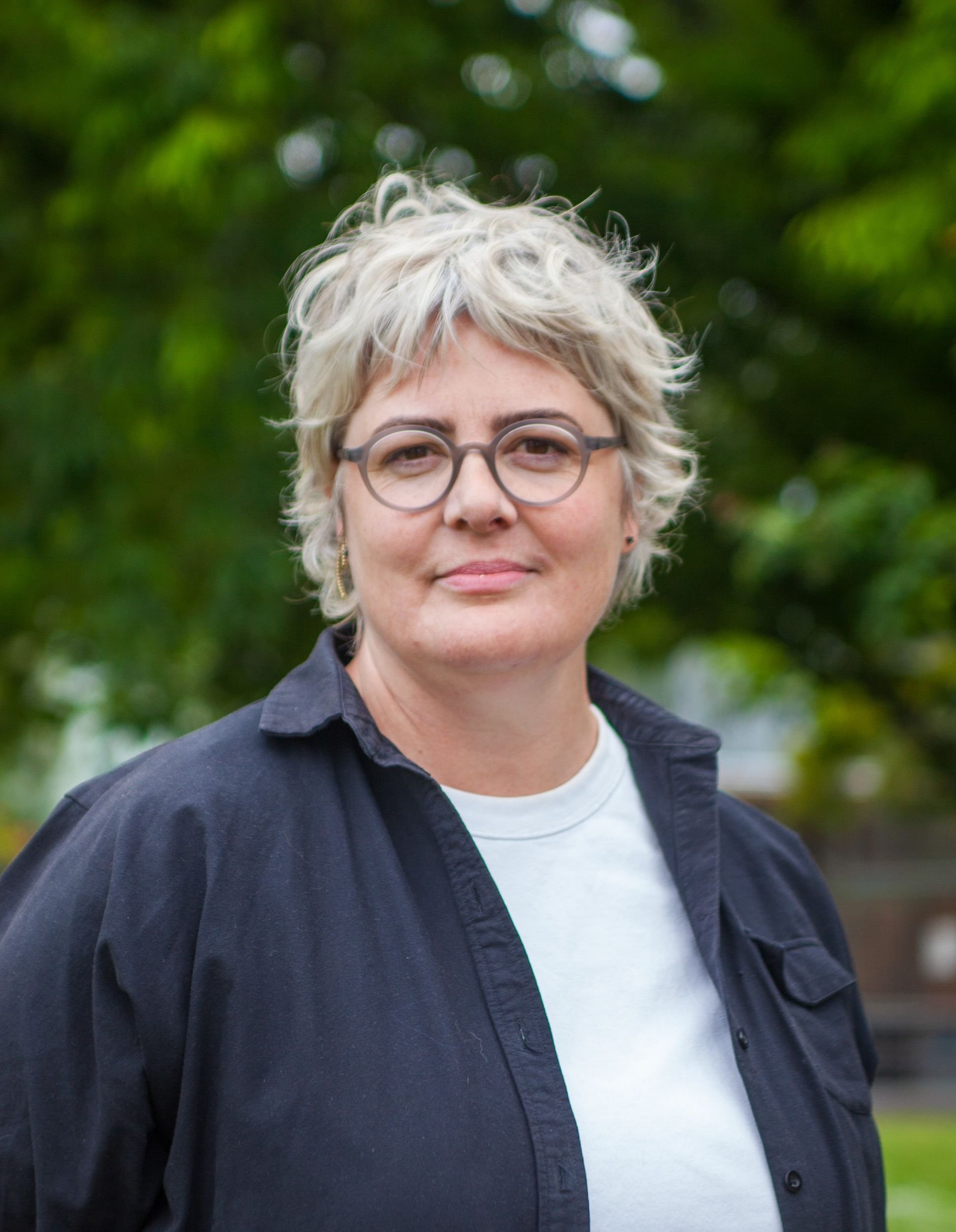 A woman with short, blond, curly hair and glasses, wearing a black shirt over a white T-shirt, standing outdoors with green trees in the background.