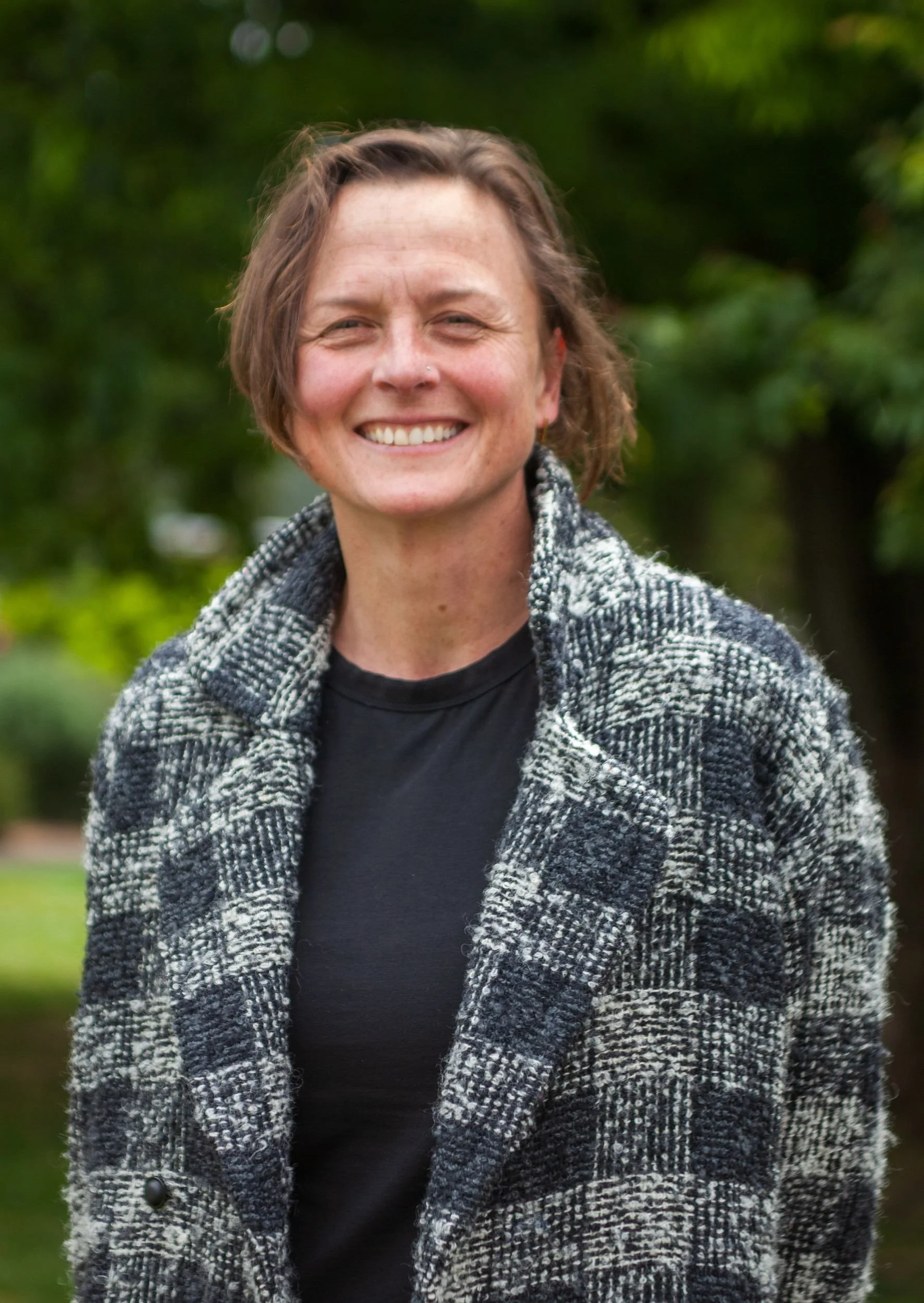 A woman smiling outdoors with a blurred green background, wearing a black shirt and a black and white plaid coat.