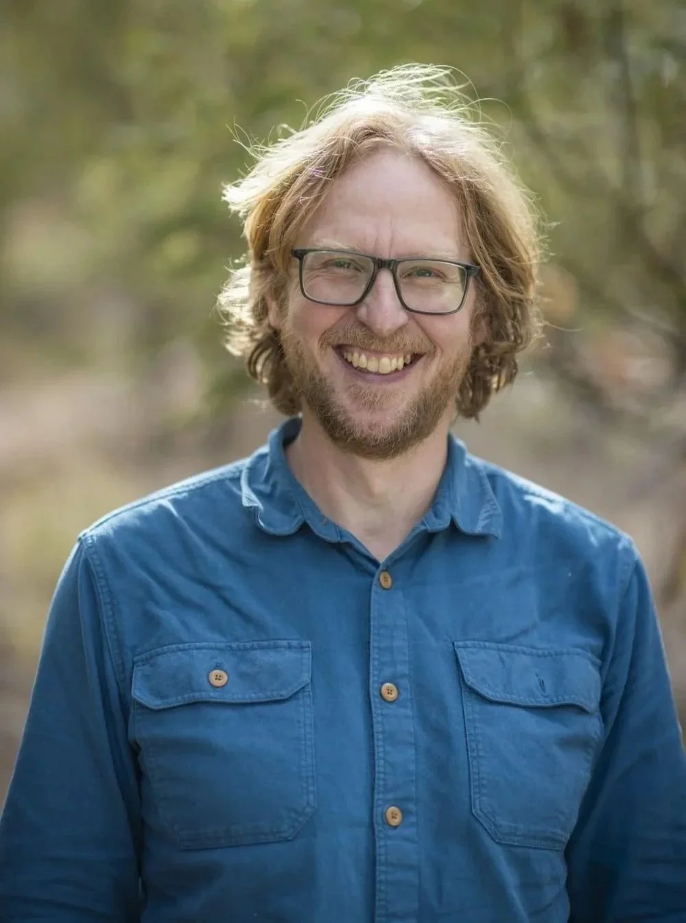 A smiling man with glasses, a beard, and long hair outdoors, wearing a blue button-up shirt.
