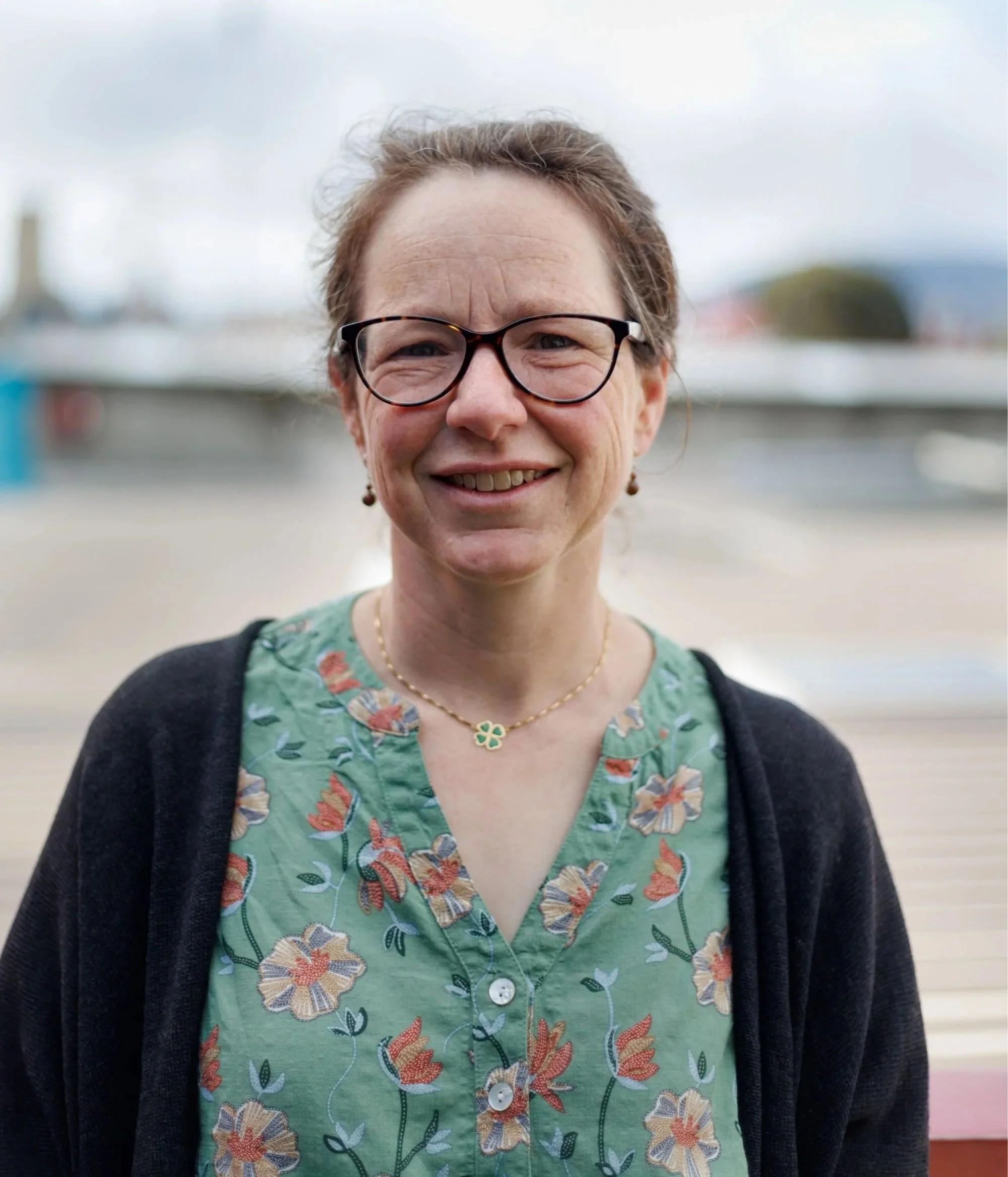A smiling woman with glasses, wearing a green floral shirt and black cardigan outdoors near water.