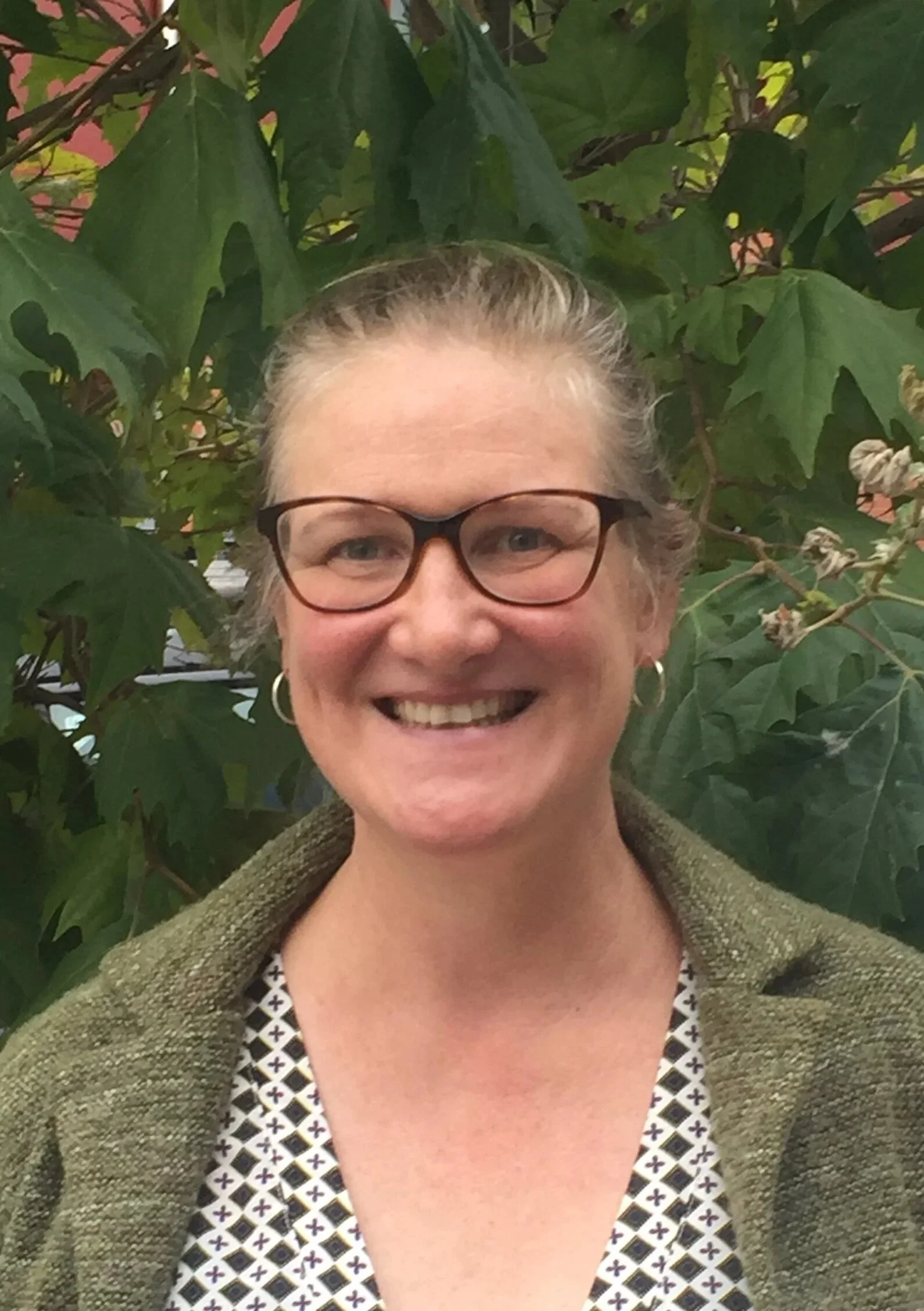A smiling woman with glasses and hoop earrings standing in front of green leafy plants.