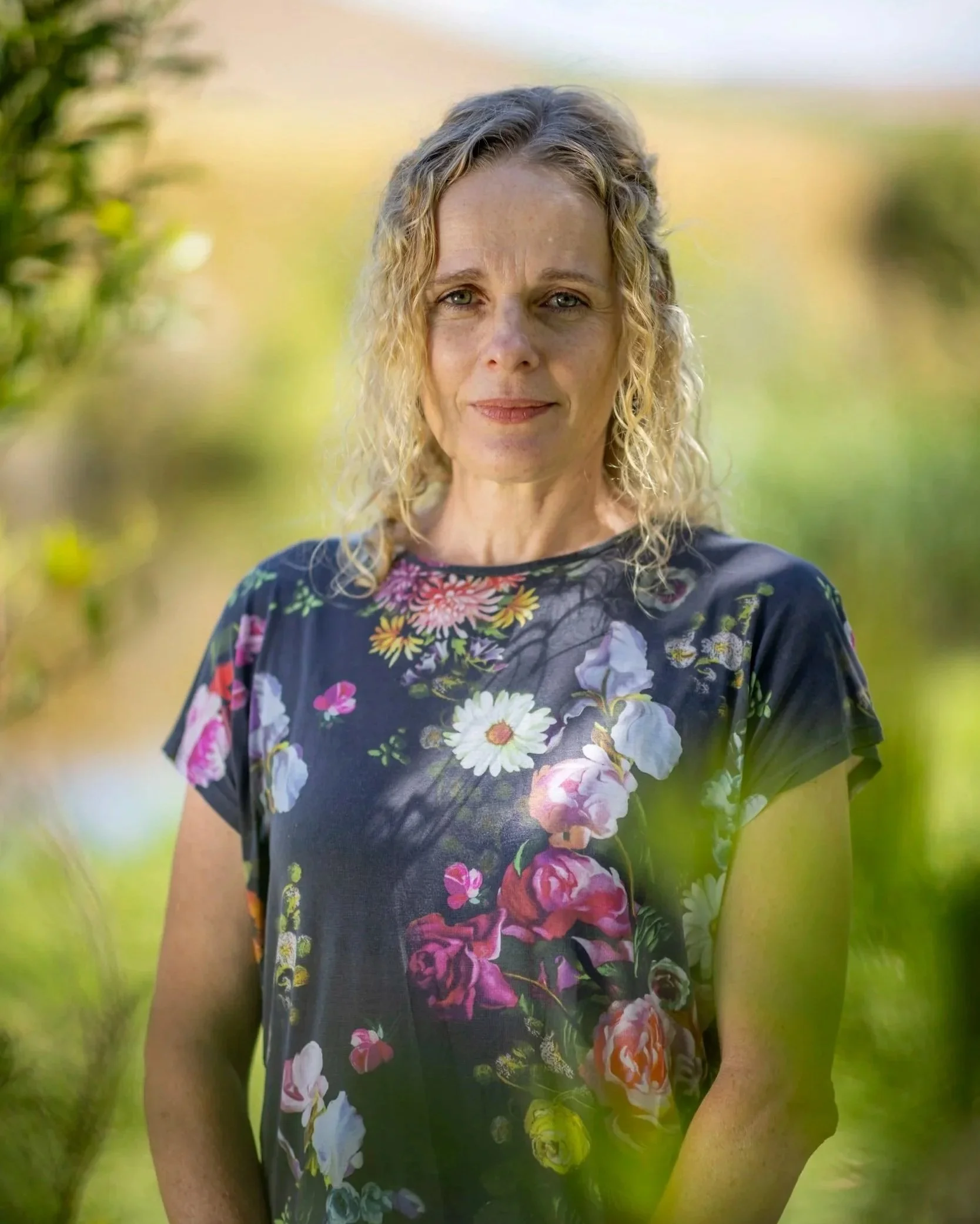 A woman with curly blonde hair smiling outdoors, wearing a black floral dress, with a blurred green natural background.