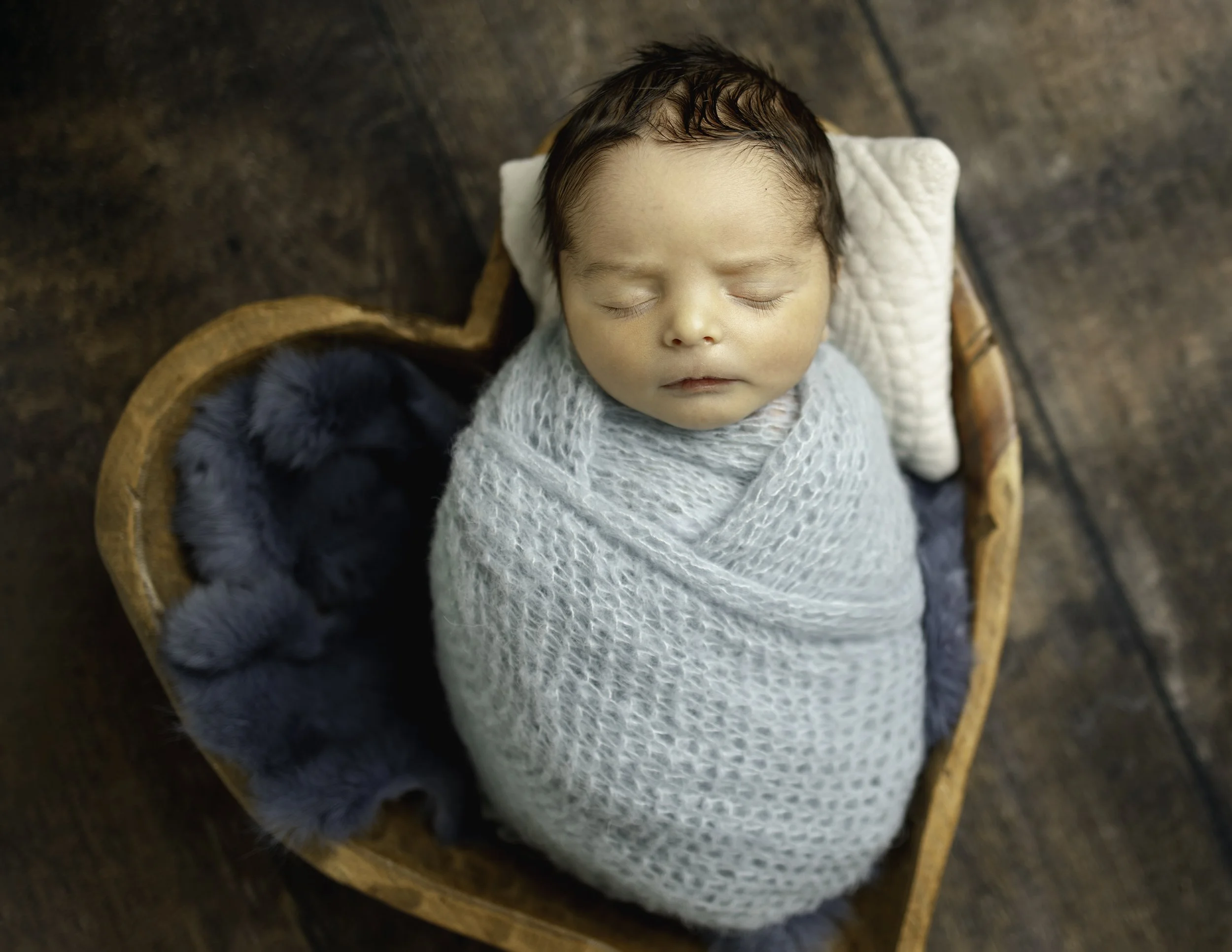 portrait of a newborn baby wrapped on a baby blue knitted wrap posed sleeping on a hwooden heart shaped bowl