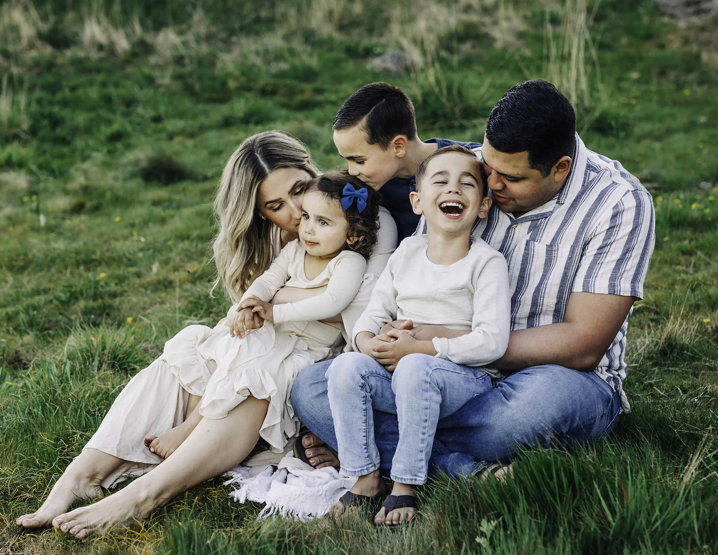 family of five seating on a meadow field with tan clothing mom is kissing little girl and dad is laughing with one of the boys in Rosario Beach WA