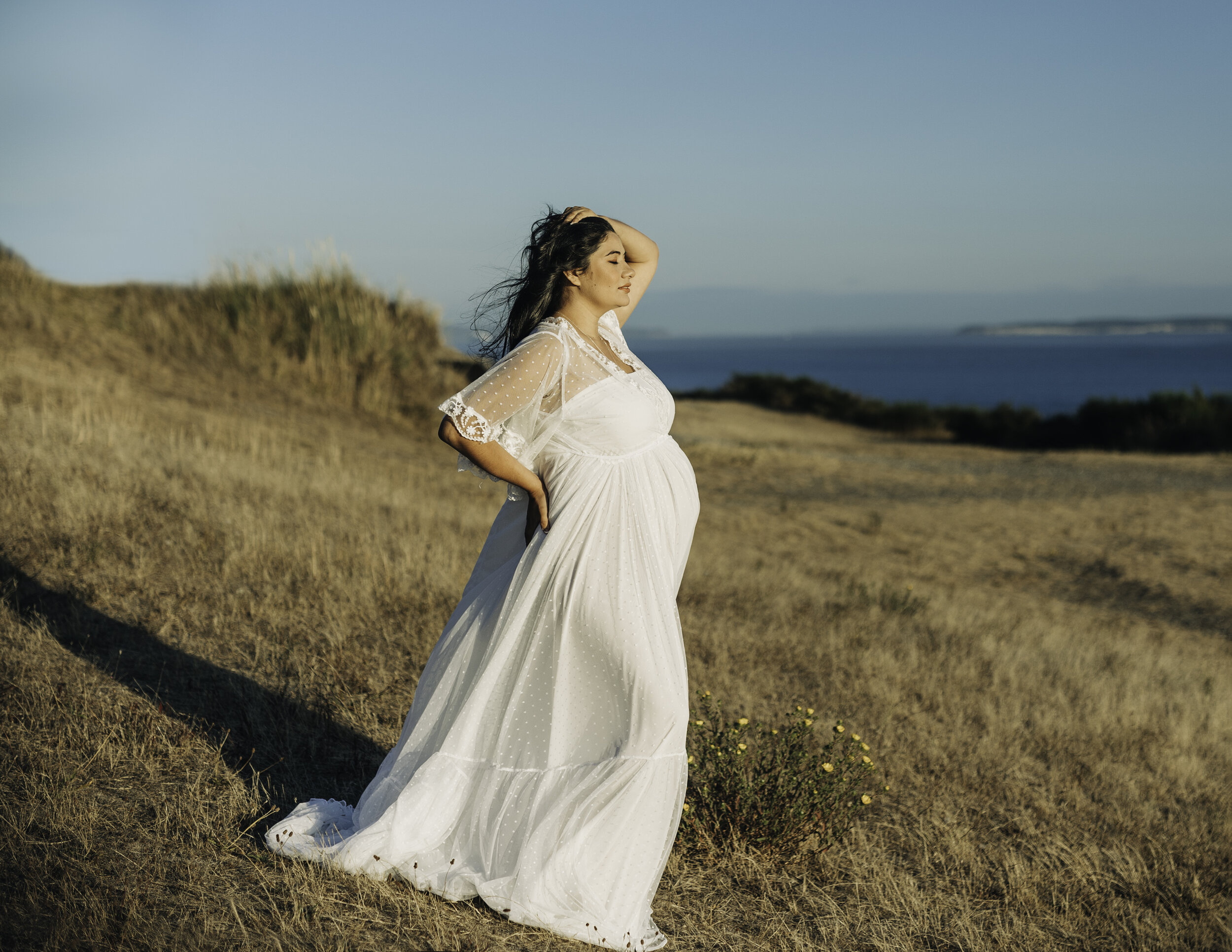 Professional maternity portrait of a pregnant woman standing in an open field wearing a white dress while holding her hips with one hand and the other one in the head, facing an ocean backdrop in Coupeville WA
