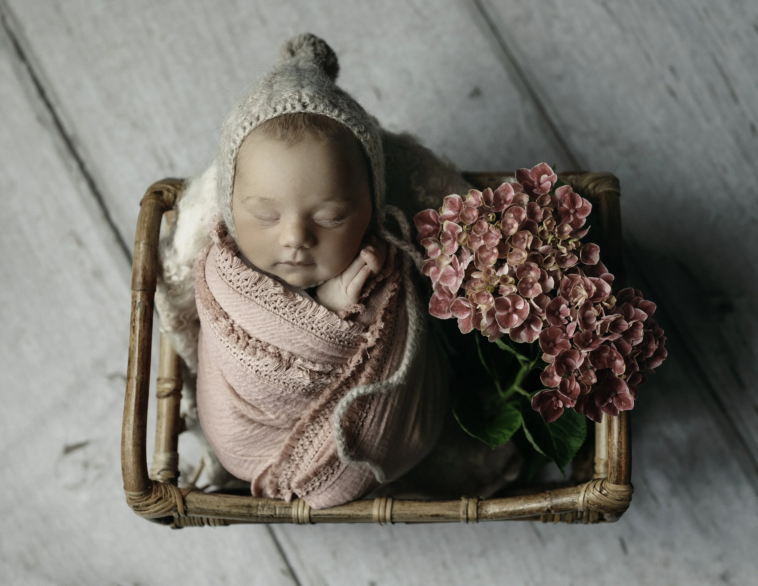 newborn photo of a baby girl wrapped in pink and posed in a bamboo basket with real flowers