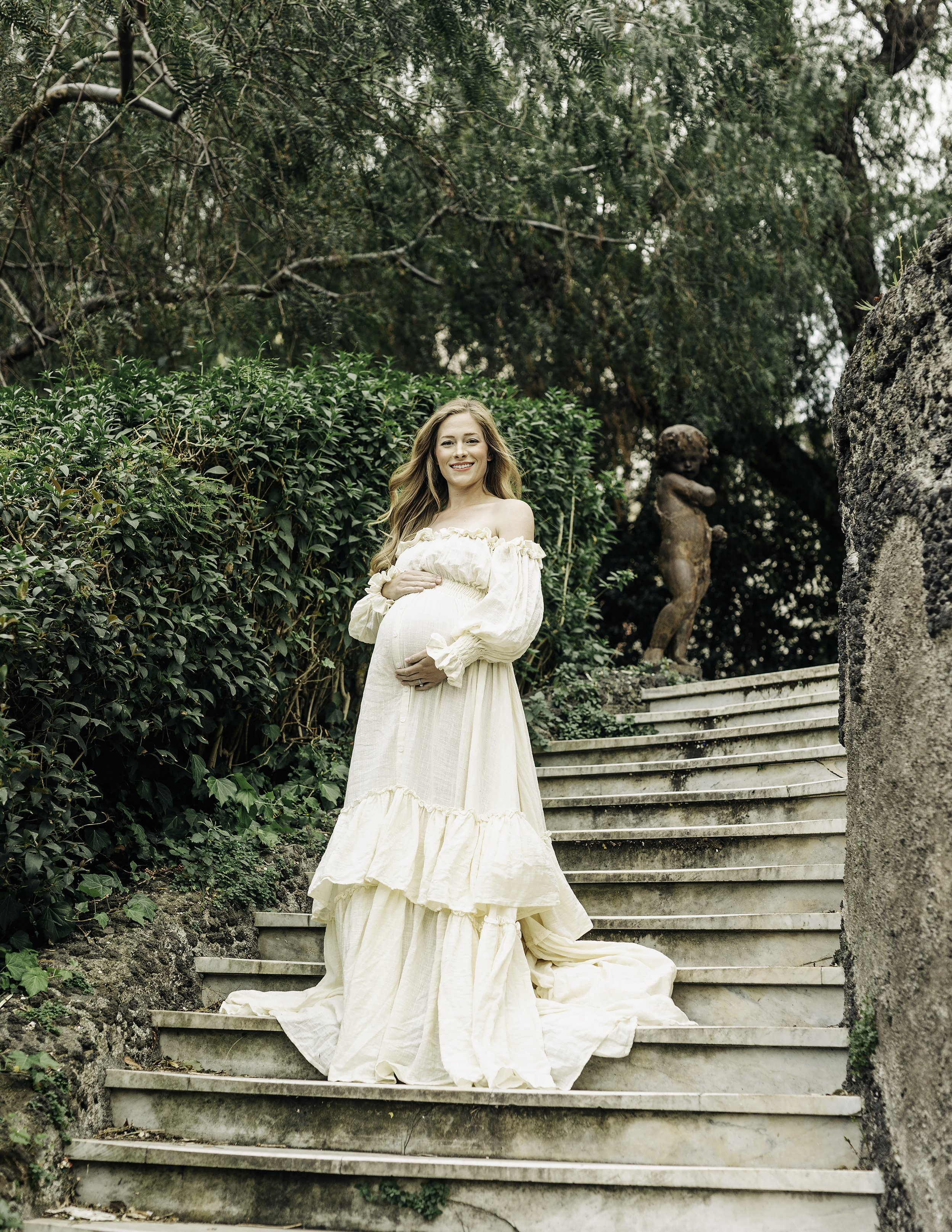 Maternity portrait of a white and blond woman wearing a light beige boho dress and holding her belly with both hands while standing on a staircase from the Bellini Gardens in Catania