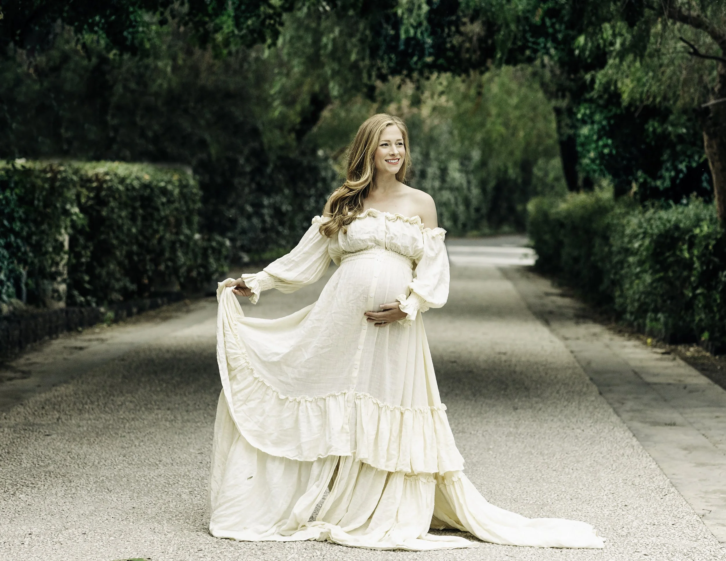 Maternity portrait of a mom to be grabbing a boho dress with one hand while holding her belly with the other hand, while looking away smiling with the Bellini Gardens in the background.