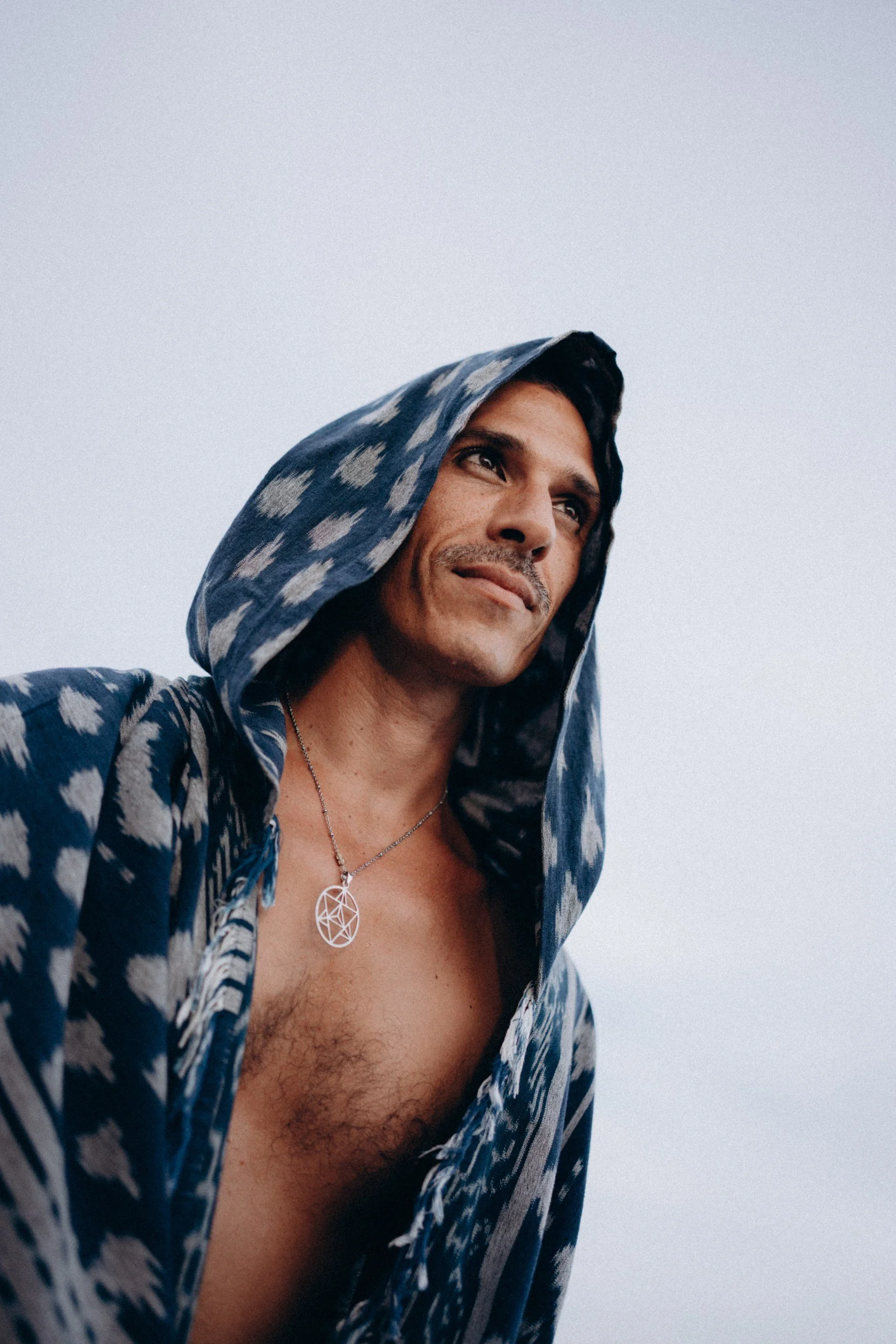 A man wearing a blue patterned hoodie with the hood over his head, looking thoughtfully into the distance, with a cloudy sky in the background, exposing part of his chest and a silver geometric necklace.