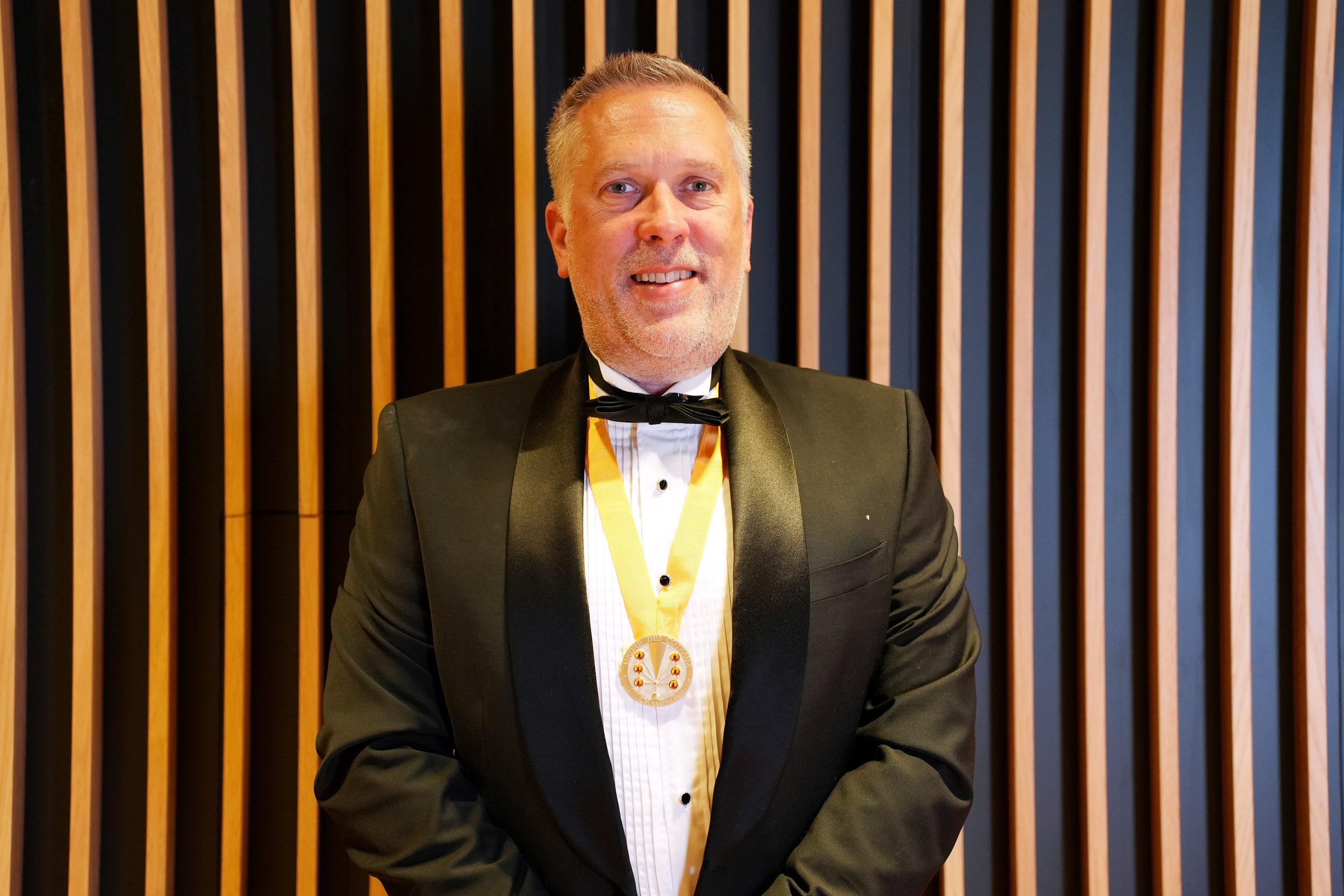 A man dressed in a black tuxedo with a black bow tie, holding a glass of champagne and wearing a green and gold medal around his neck, at a formal event or celebration.