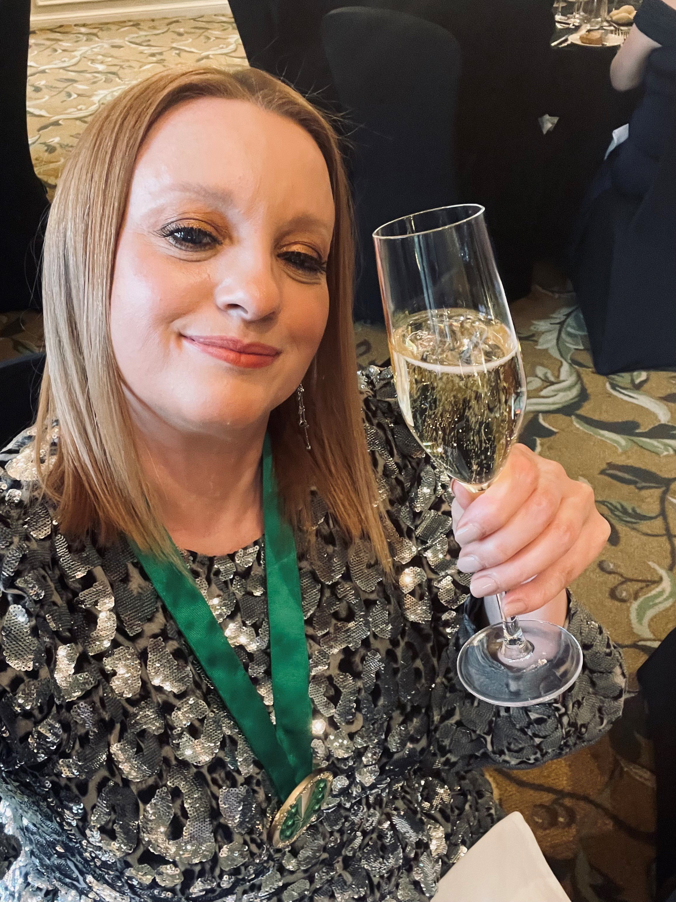 A woman with shoulder-length hair and makeup, smiling while holding a glass of champagne at a formal event.