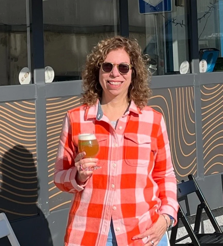 Woman with curly hair, wearing sunglasses and a bright red and pink plaid shirt, holding a beer glass outside on a sunny day.