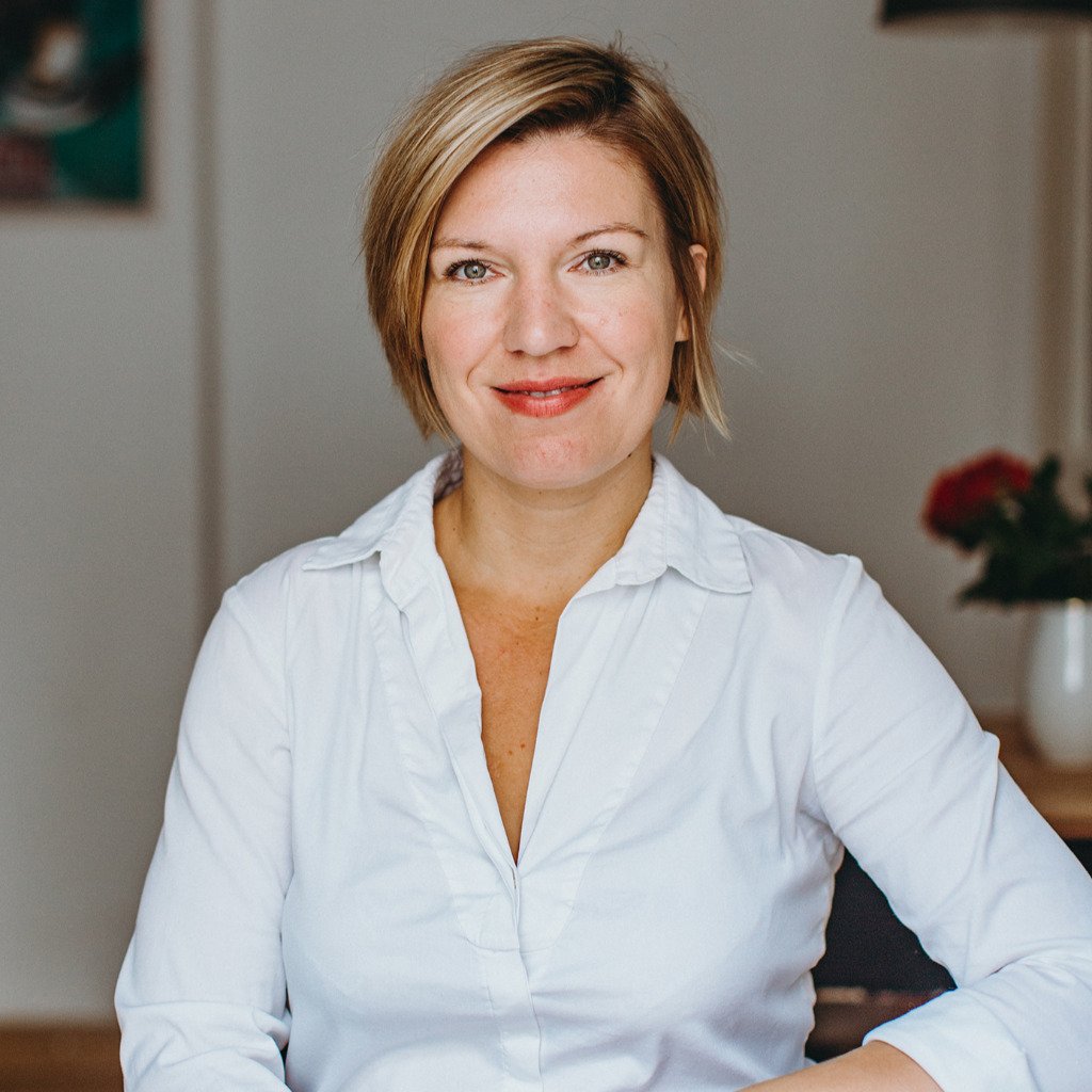 A woman with short, light brown hair wearing a white button-up shirt, smiling indoors with a blurred background that includes a white vase with red flowers.