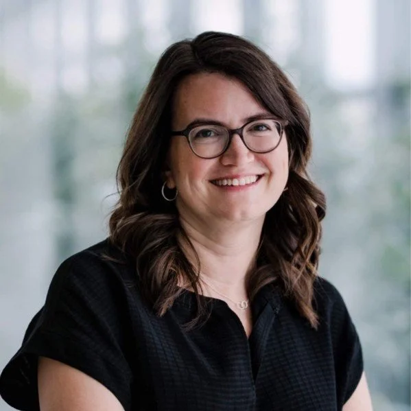 Young woman with shoulder-length brown hair, glasses, and hoop earrings, smiling in a professional headshot with a blurred window background.
