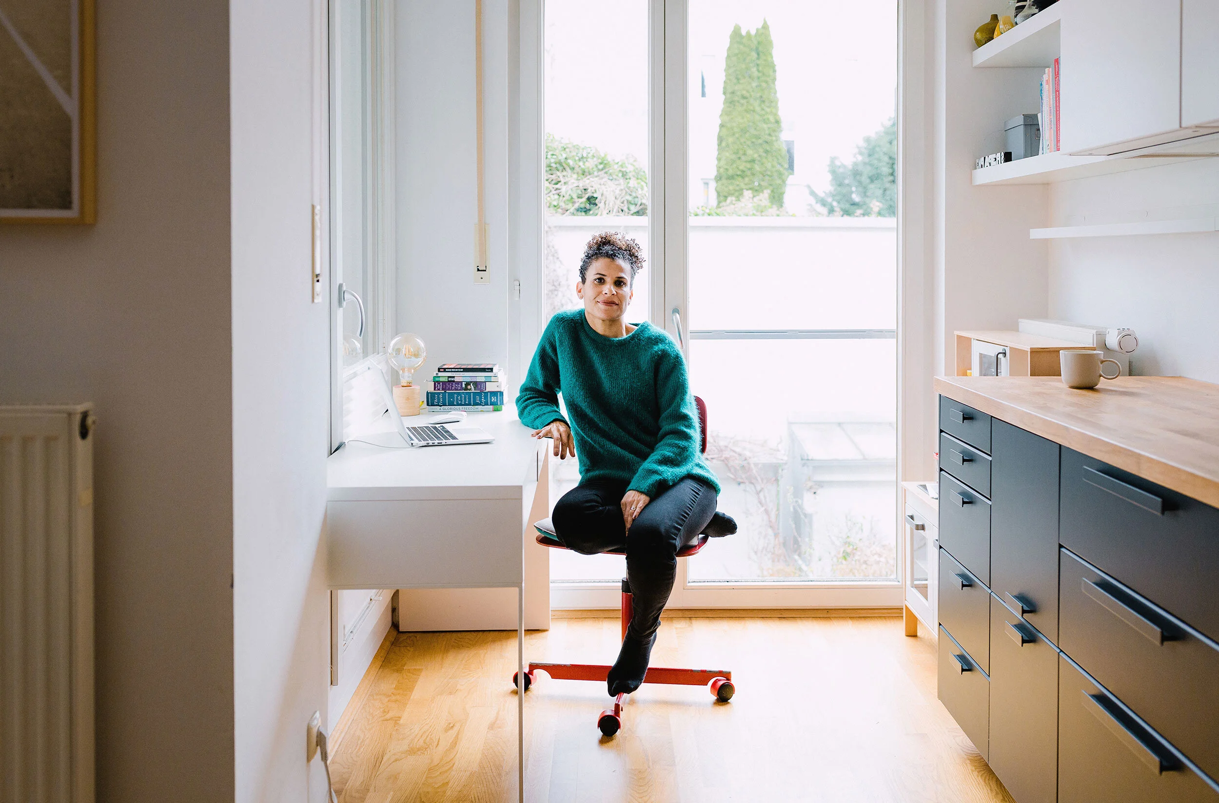 A woman sitting on a red rolling office chair in a bright kitchen with large windows behind her, wearing a teal sweater and black pants.