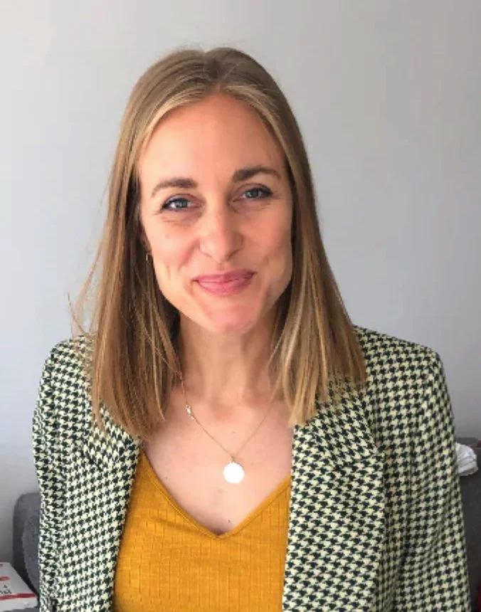 A woman with shoulder-length blonde hair, smiling, wearing a houndstooth blazer over a yellow top, standing against a plain light gray wall.