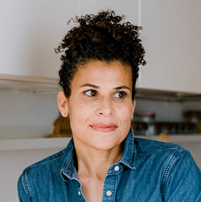 A woman with short, curly hair wearing a denim shirt, looking to the side indoors.