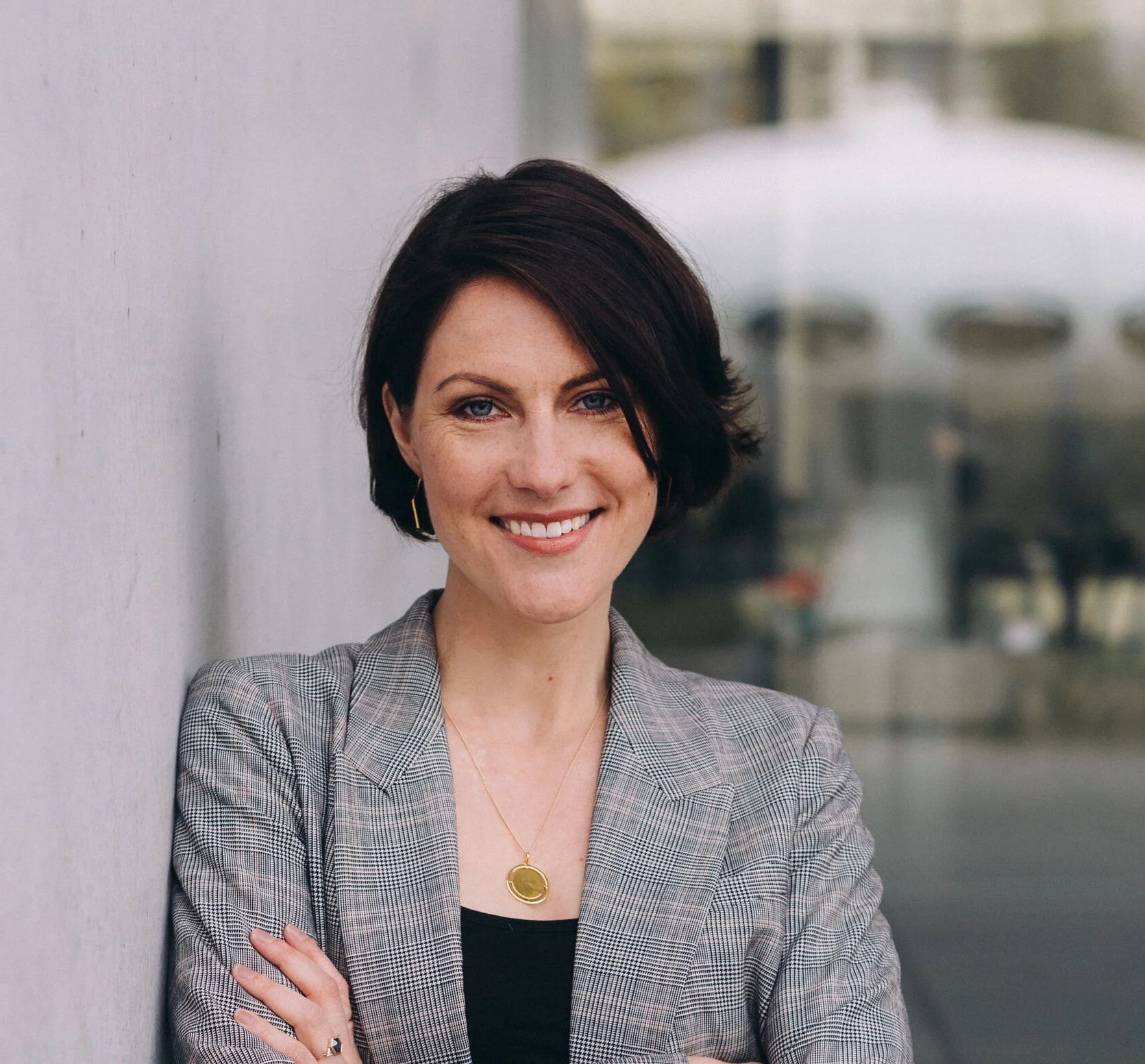 A woman with short dark hair smiling, wearing a checkered blazer and gold jewelry, standing outdoors in front of a light-colored wall with a blurred background.